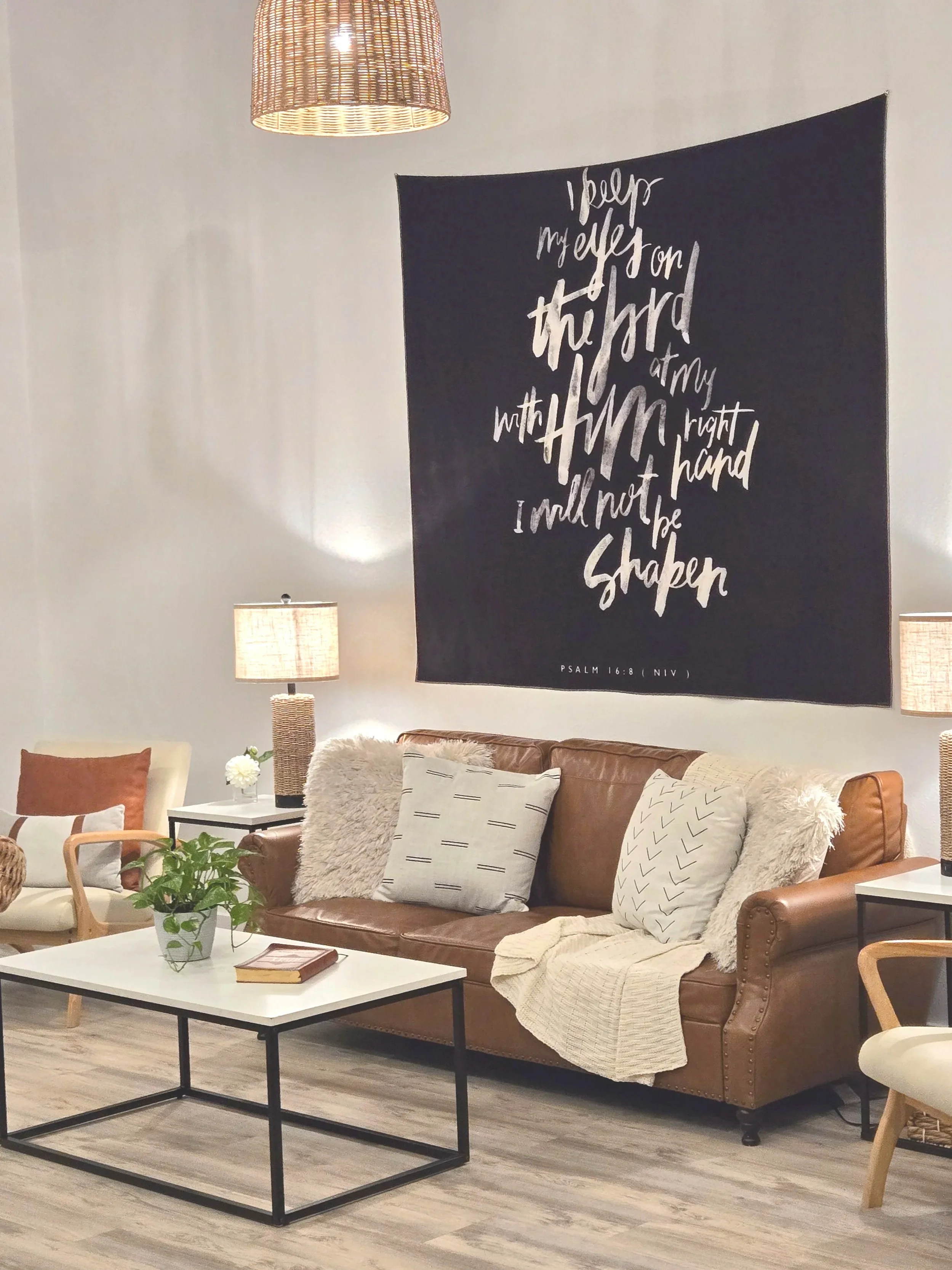 Living room with a brown leather sofa decorated with various pillows and a throw blanket, a white coffee table with a potted plant and book, two side tables with lamps, a tapestry with a quote on the wall, and a woven light fixture hanging from the c