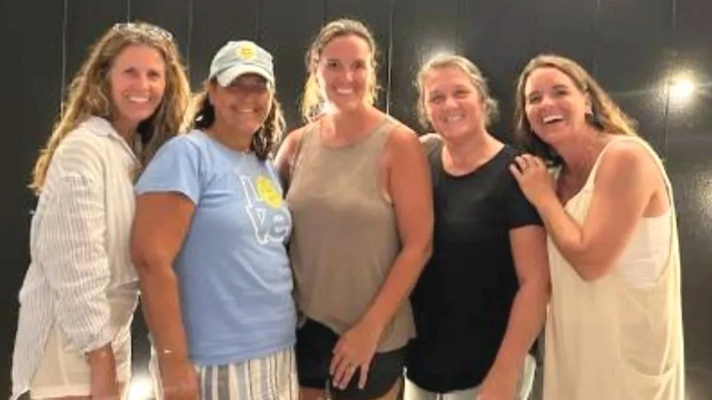 Five women smiling and standing close together in a casual setting.