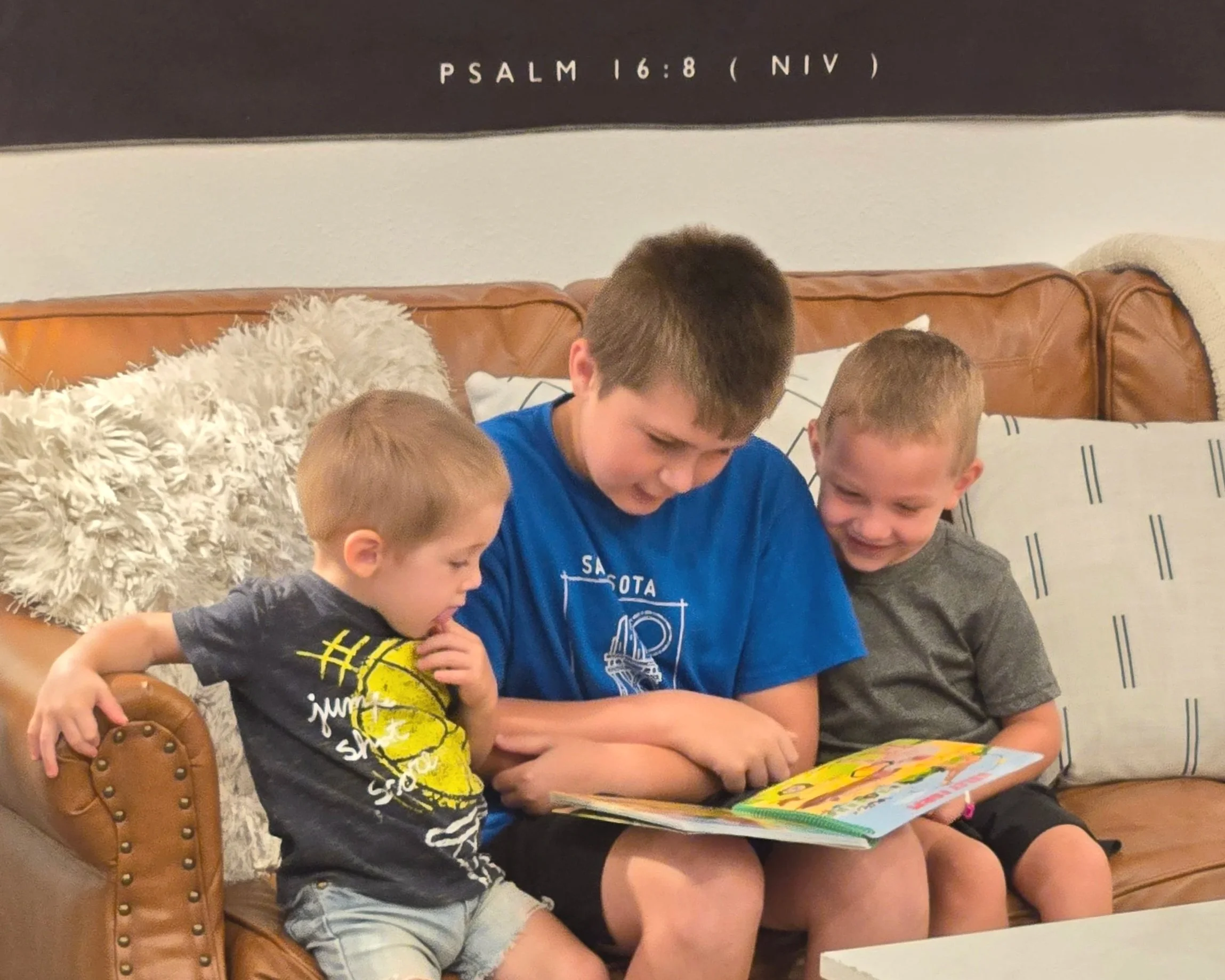 Three boys sitting on a brown leather couch, reading a colorful children's book together, with pillows and a blanket behind them.