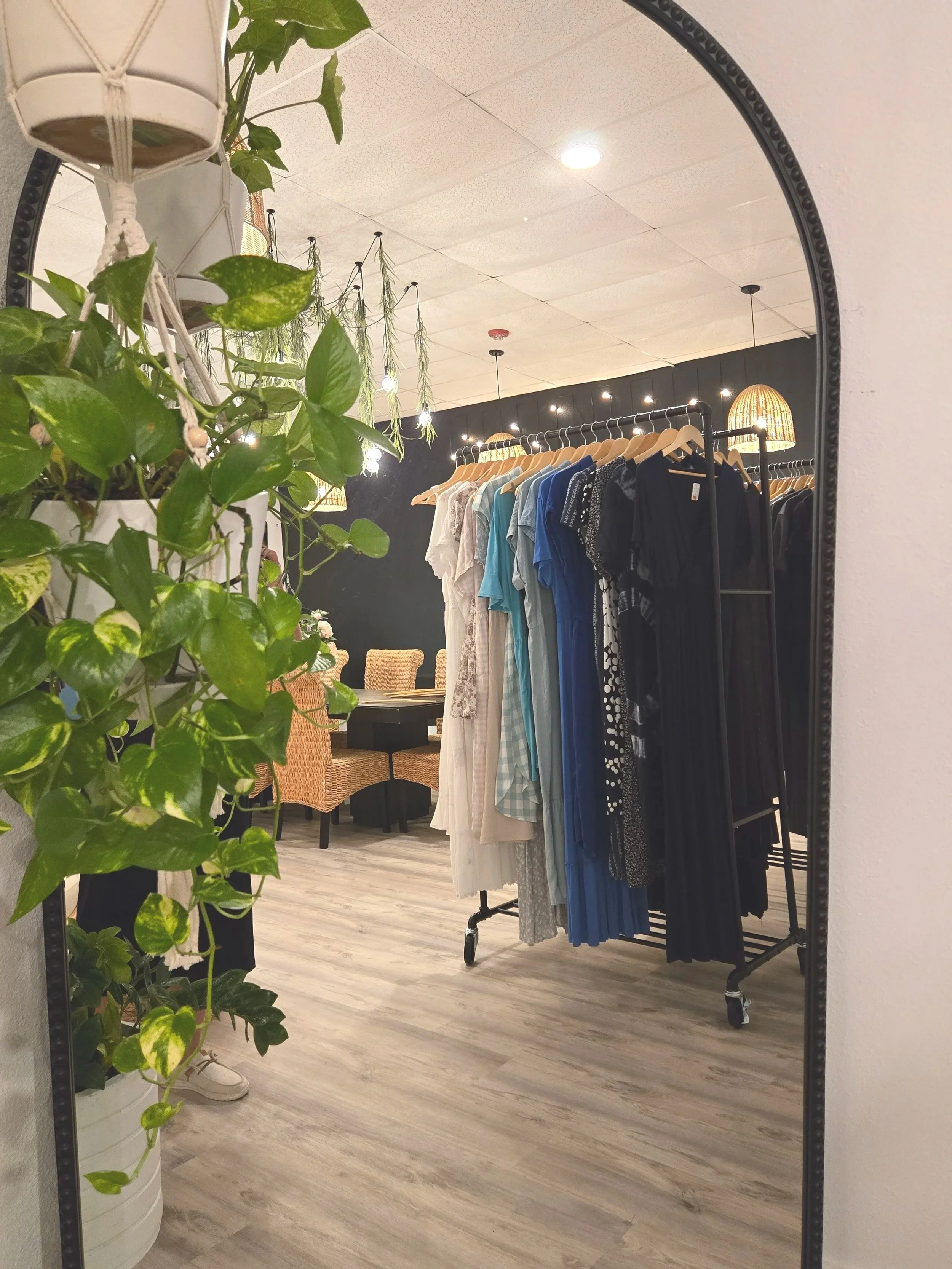 Clothing store with rack of dresses and tops, plants, and seating area reflected in a mirror.
