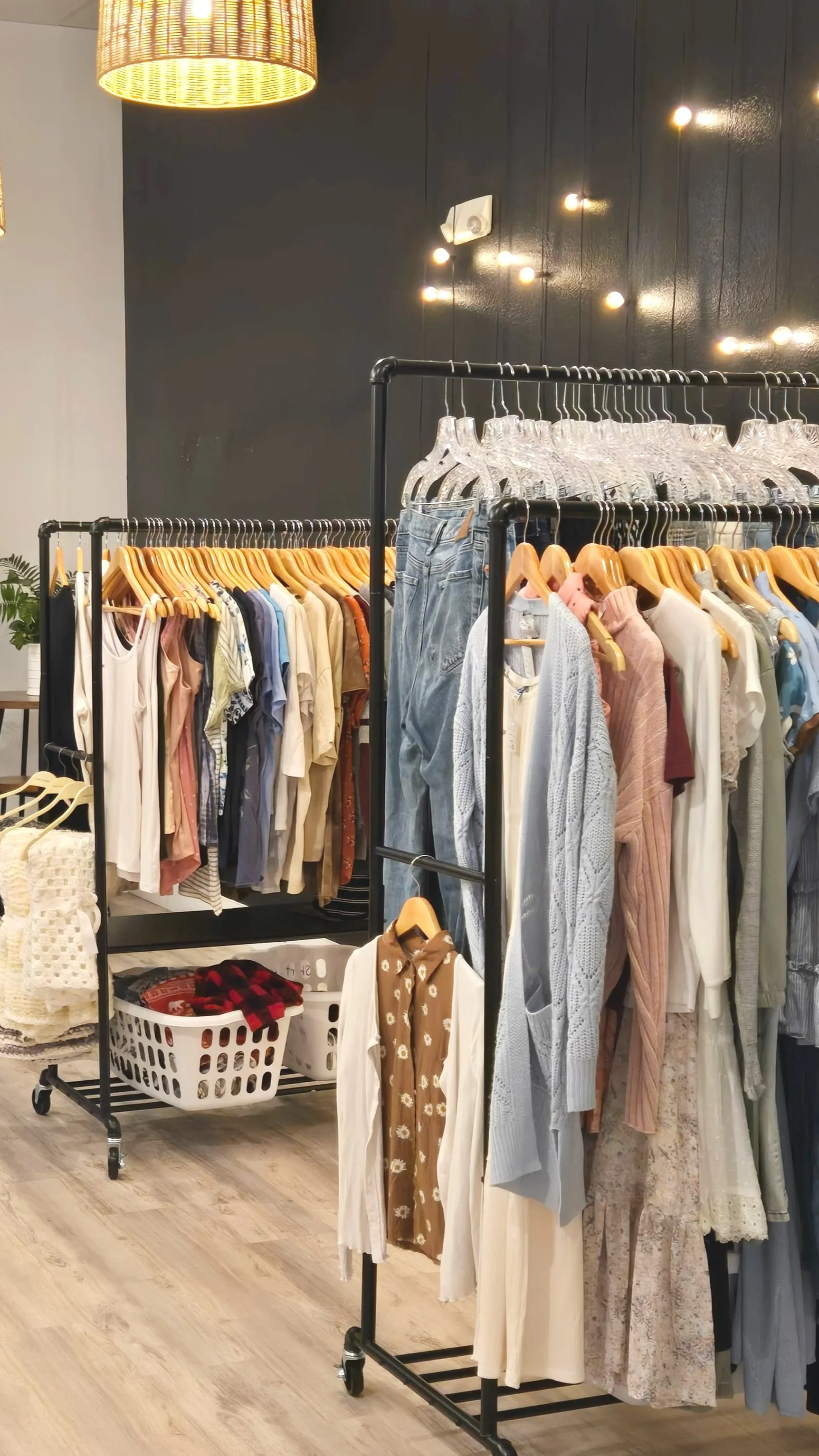 Clothing rack with various casual clothes hanging on wooden hangers, in a boutique store with wooden flooring, dark wall, and string lights