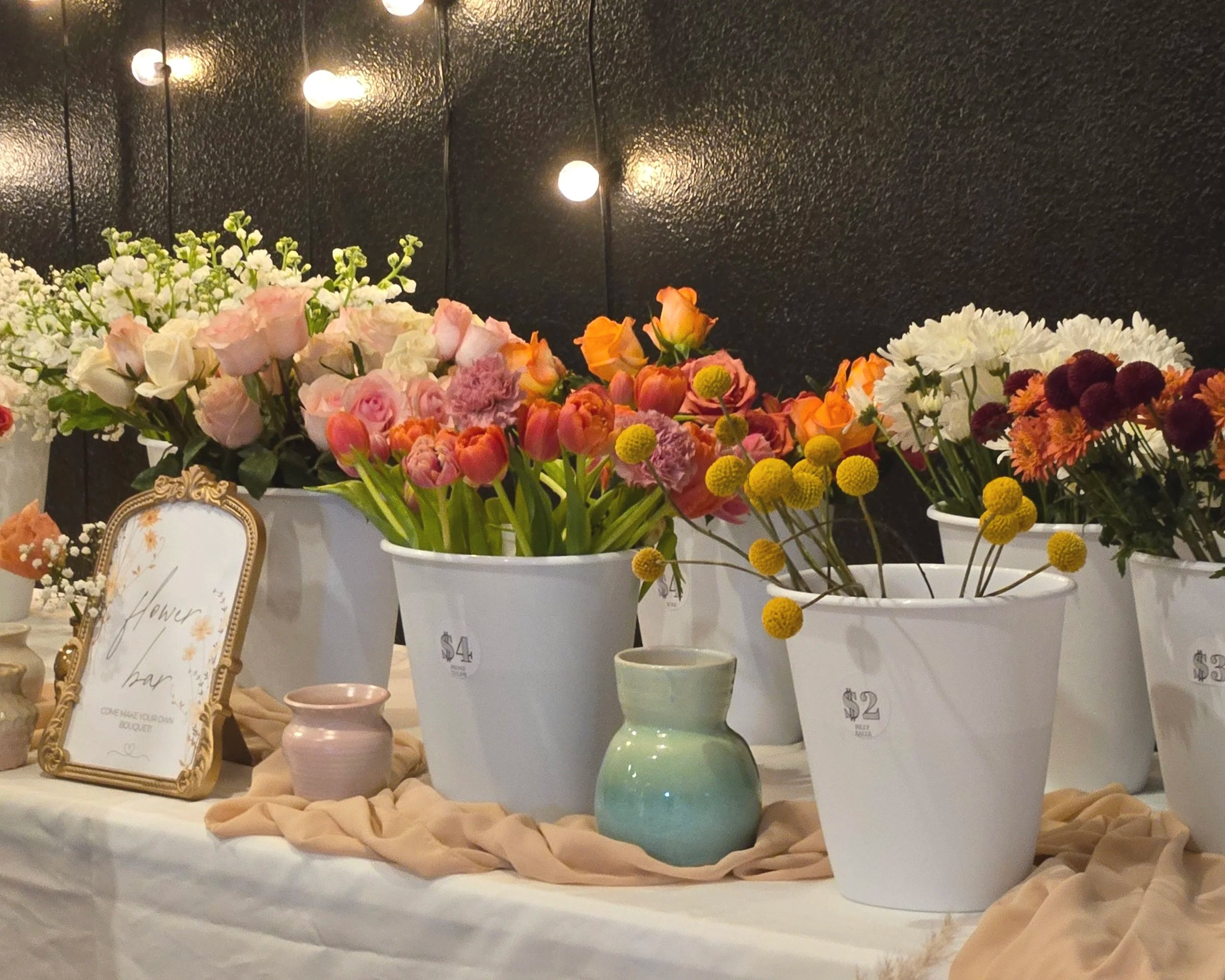 Display of colorful flower bouquets in white pots at a flower bar, with a framed sign reading 'Flower Bar' on a table draped with beige fabric and decorative vases.