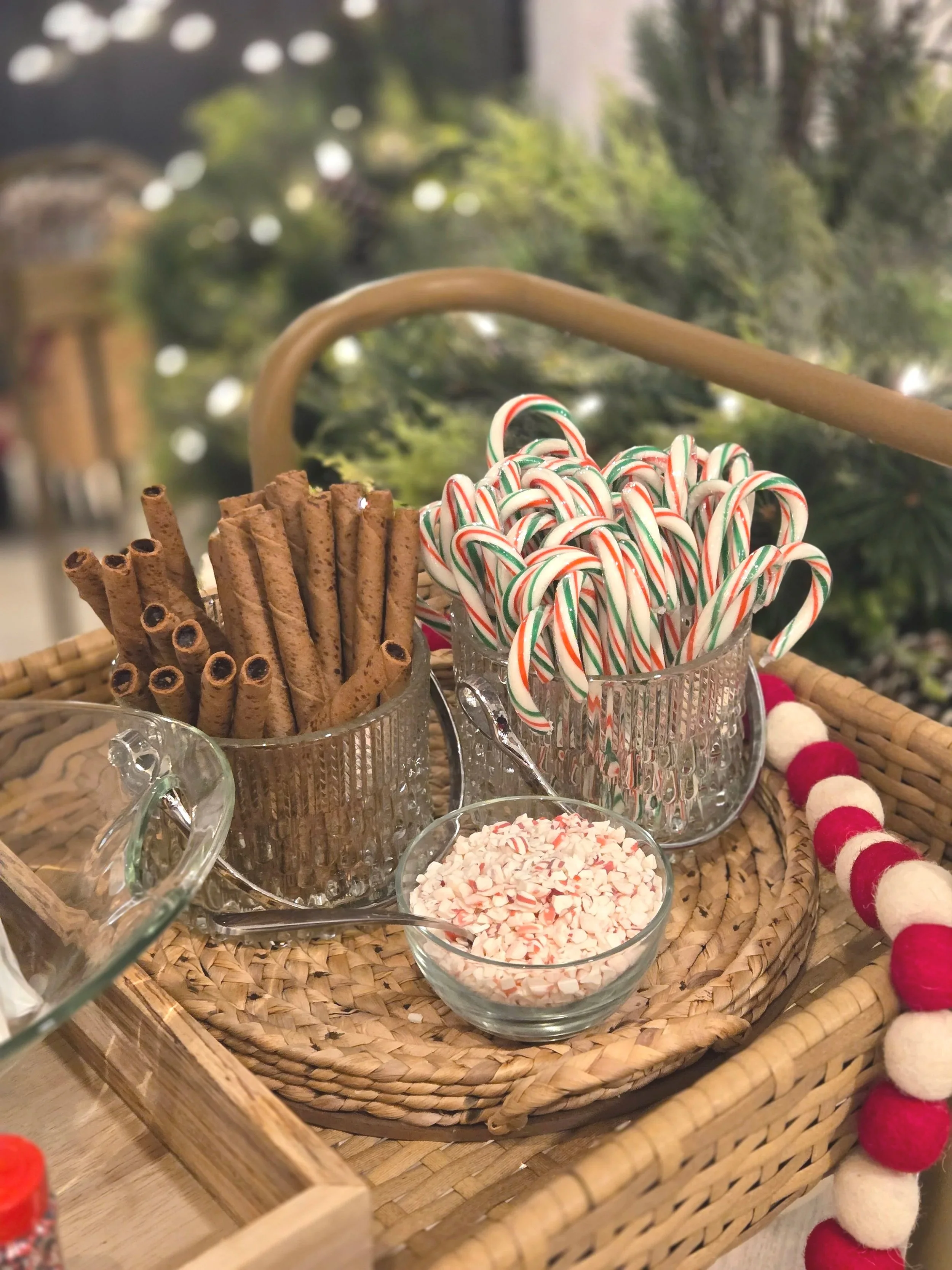 A wicker basket containing glass jars filled with candy canes, cinnamon sticks, and crushed peppermint, with a bowl of crushed peppermint on a holiday-themed tray.