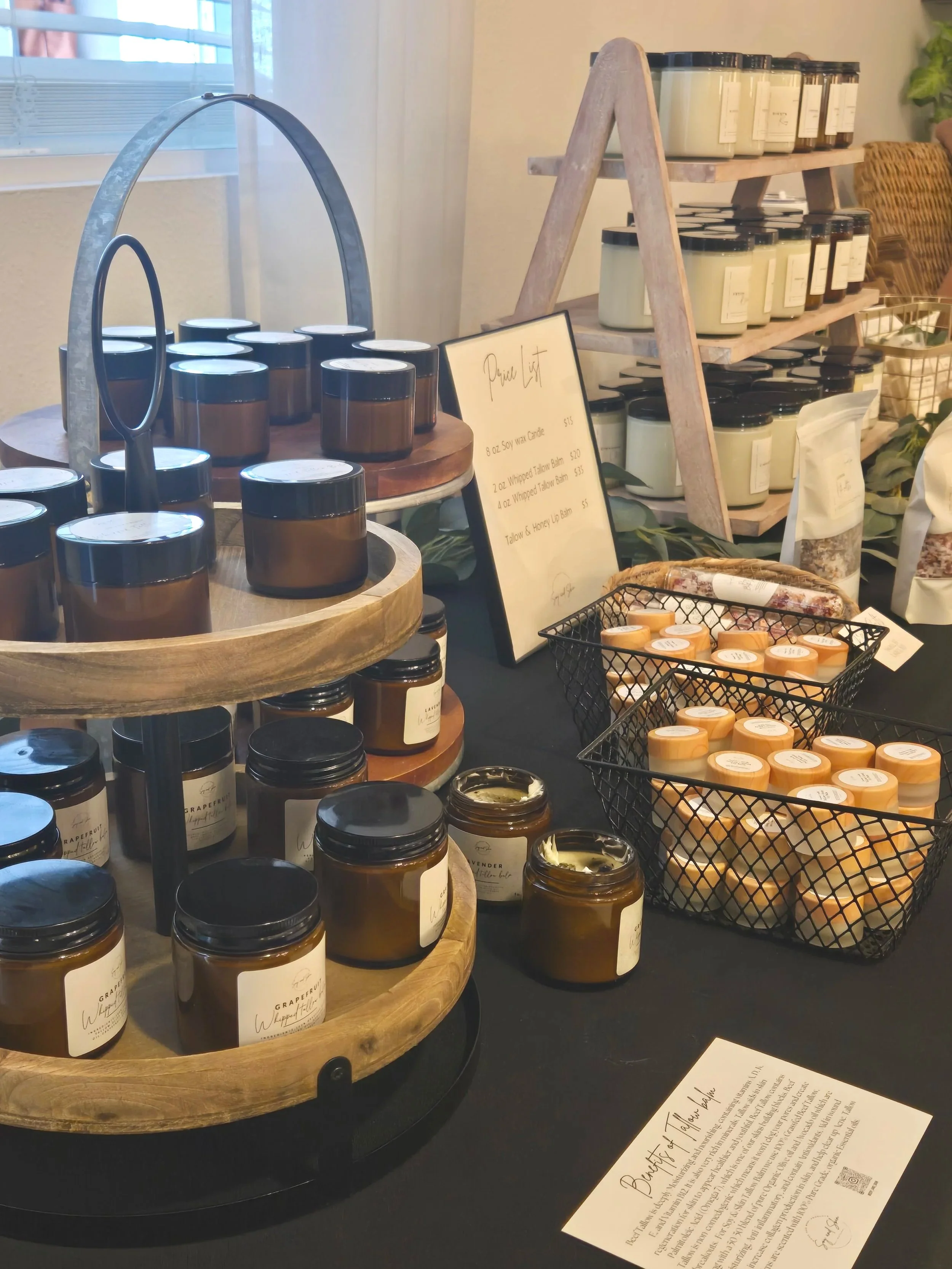 Display of various candles and wax melts at a market booth. Several amber jars with black lids, a wire basket filled with small round wax melts, and a framed menu listing different candle options. Shelves in the background hold additional candles or 