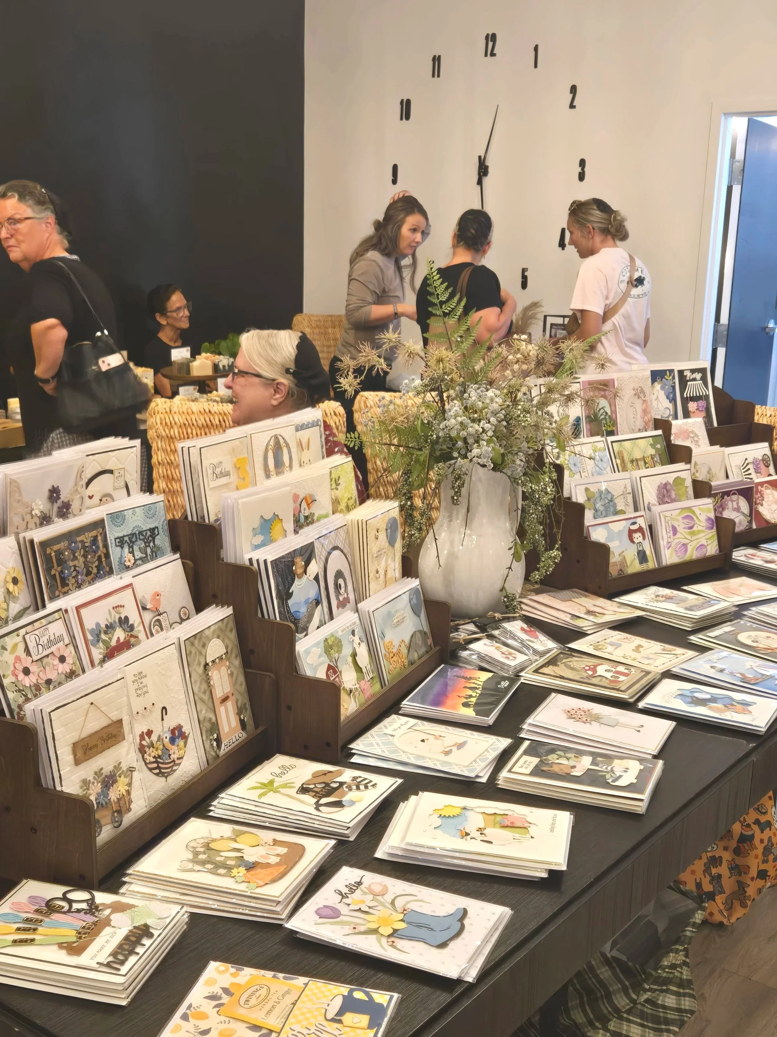 A tabletop display of greeting cards, with a large flower vase holding white and green flowers at the center. In the background, several people are browsing and chatting, with some seated and others standing, against a wall with a large clock showing