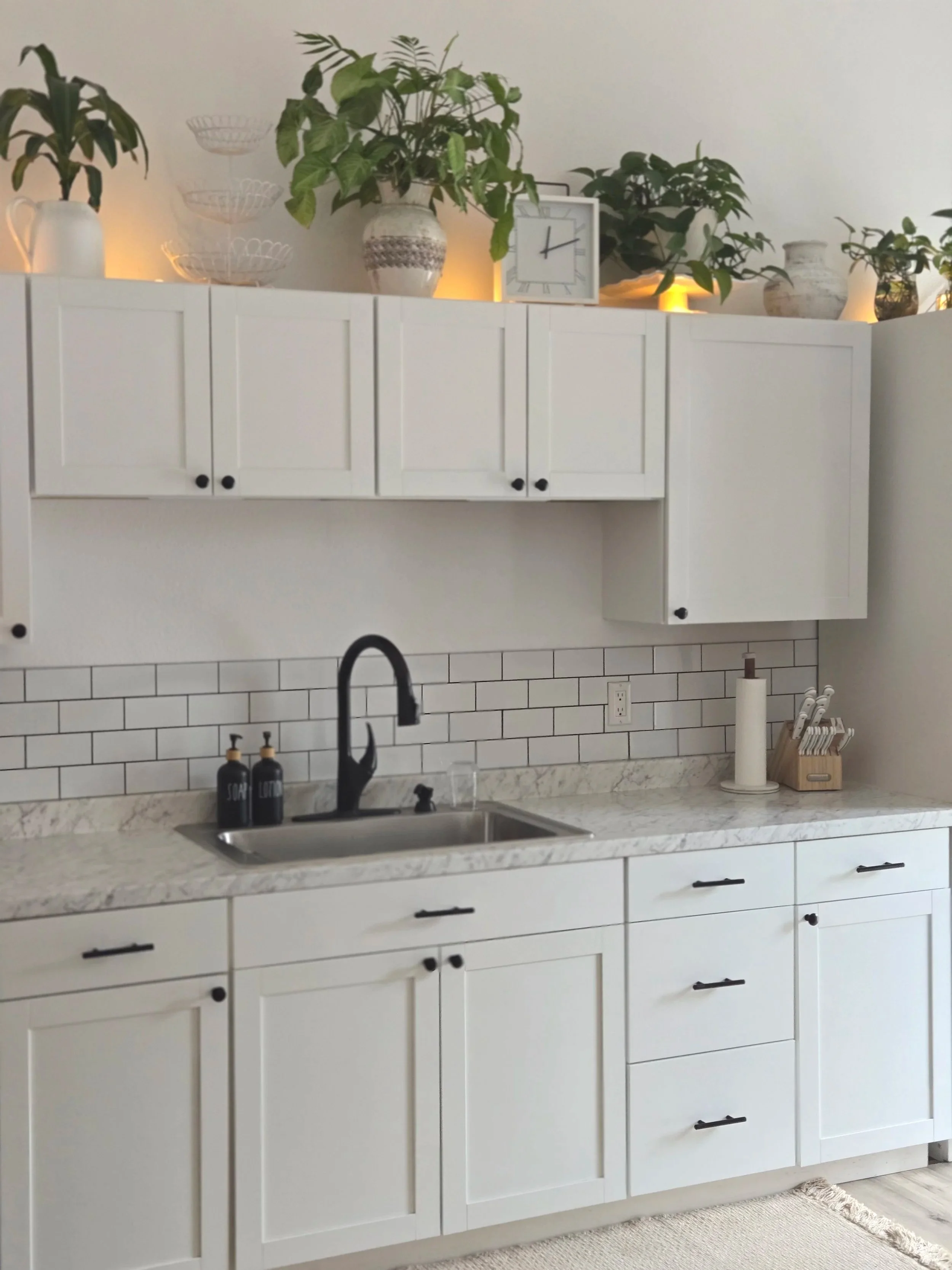 White kitchen with black handles, marble countertop, black faucet, small plants and decorative items on upper cabinets, and kitchen accessories on the counter.