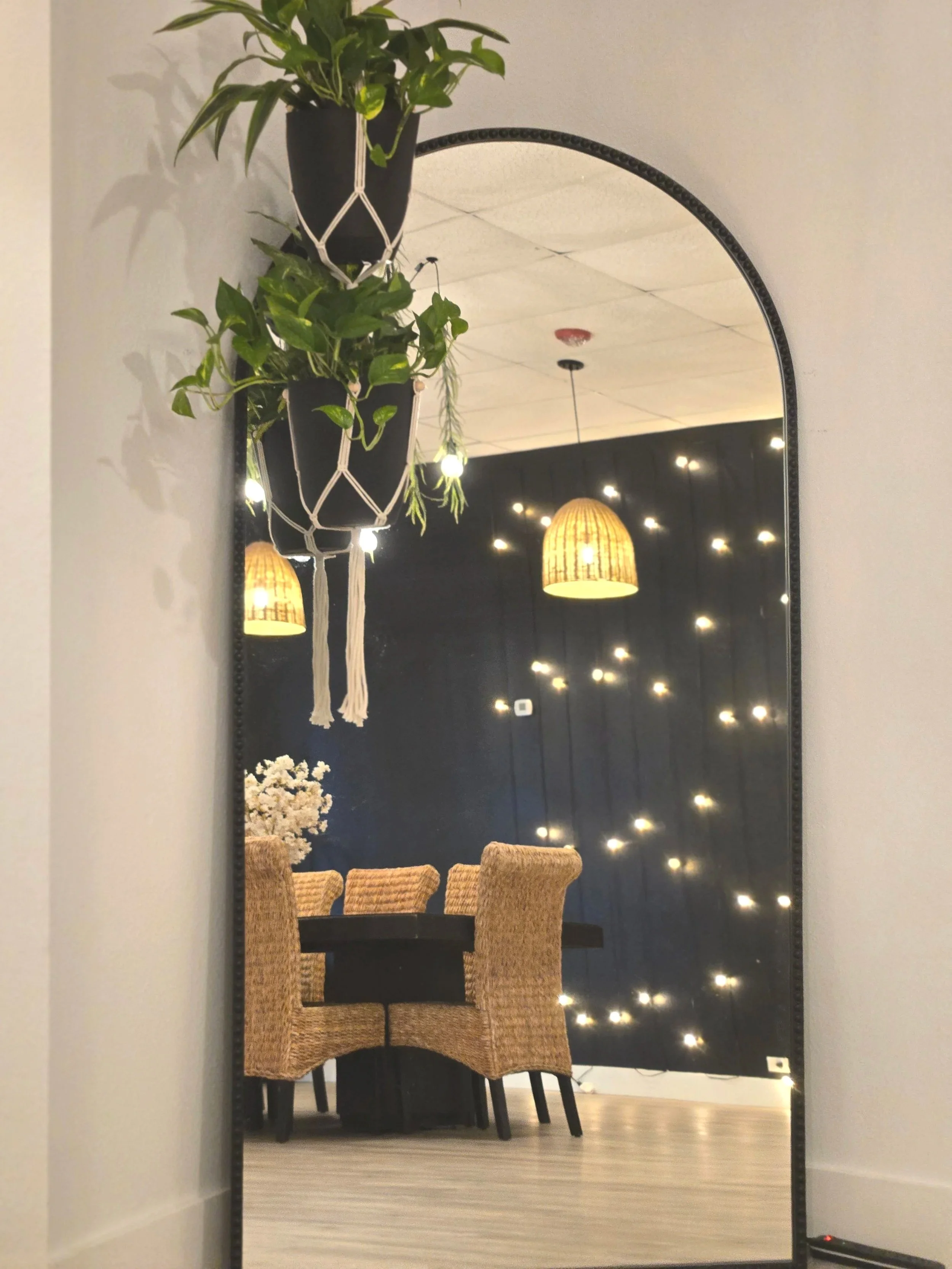 A mirror reflecting a dining area with a dark background wall decorated with string lights, a table with four rattan chairs, and hanging woven pendant lights.