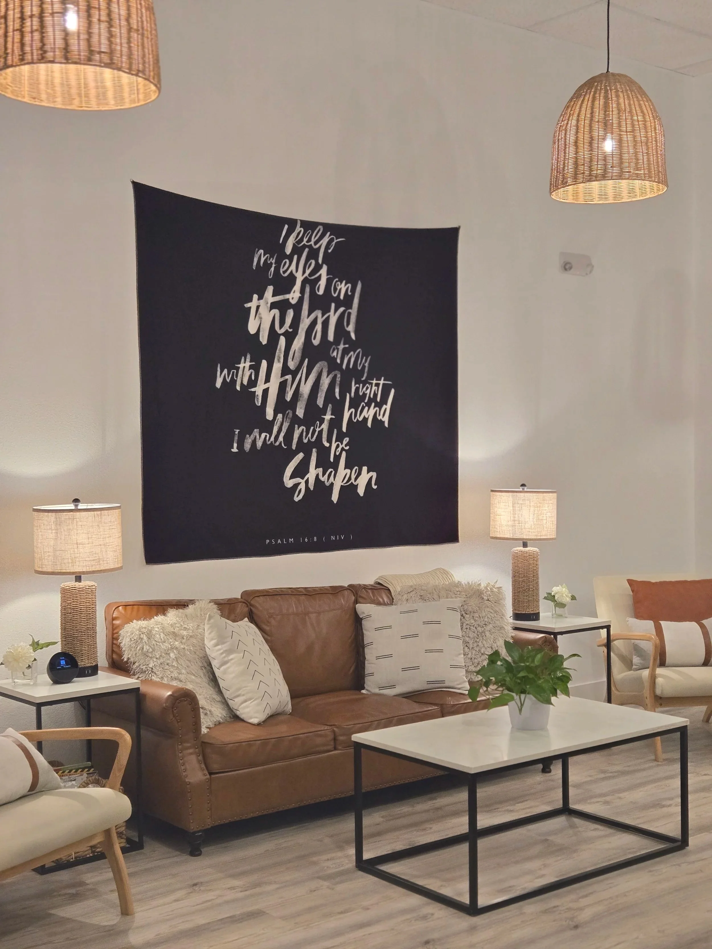 Living room with a brown leather sofa and various pillows, a white coffee table with a potted plant, side tables with lamps, and wall hanging with a biblical verse.