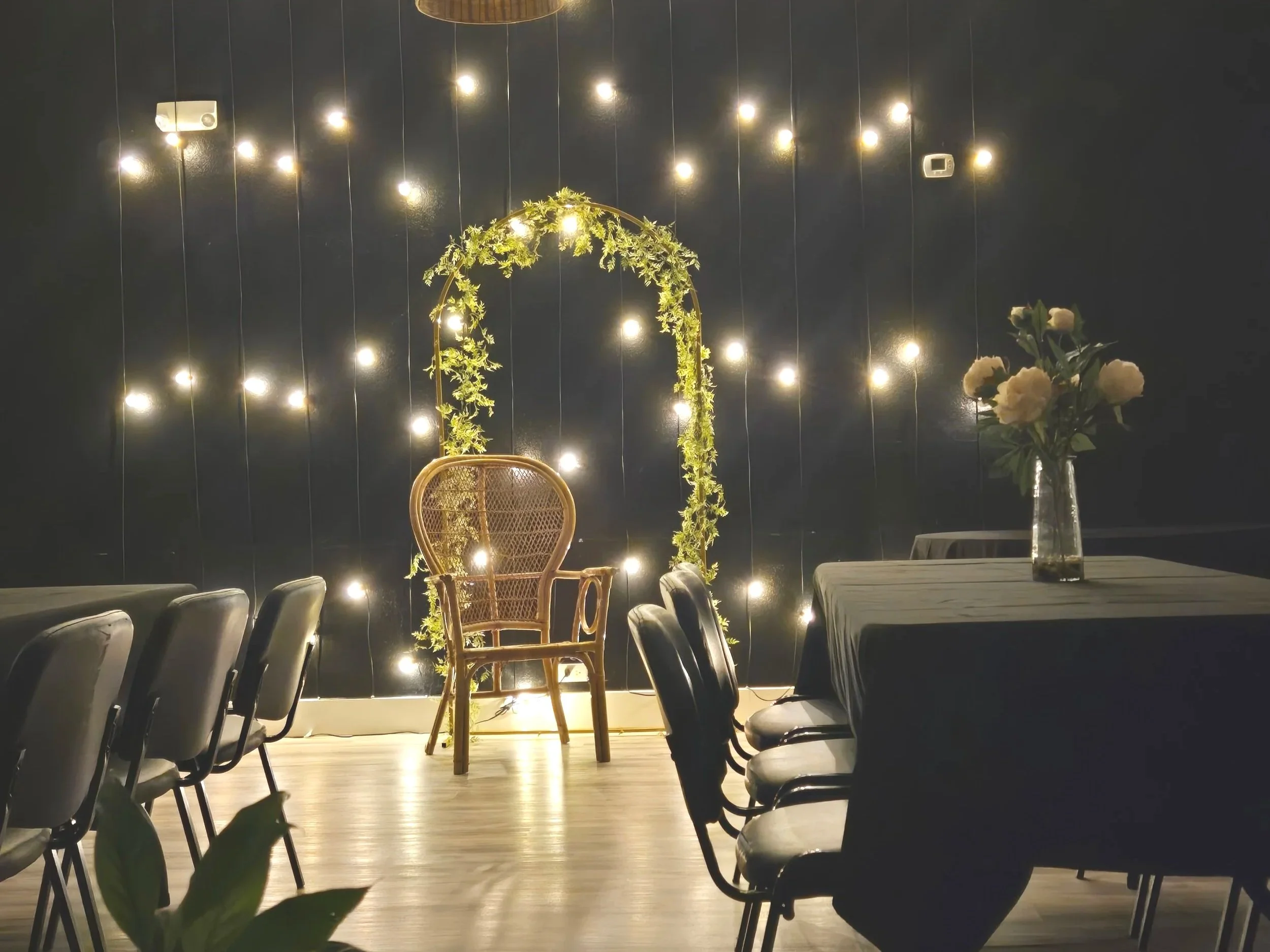 Decorative setup with a rattan chair under an arch decorated with greenery, illuminated by hanging string lights on a dark wall, adjacent to a table with a vase of white flowers.