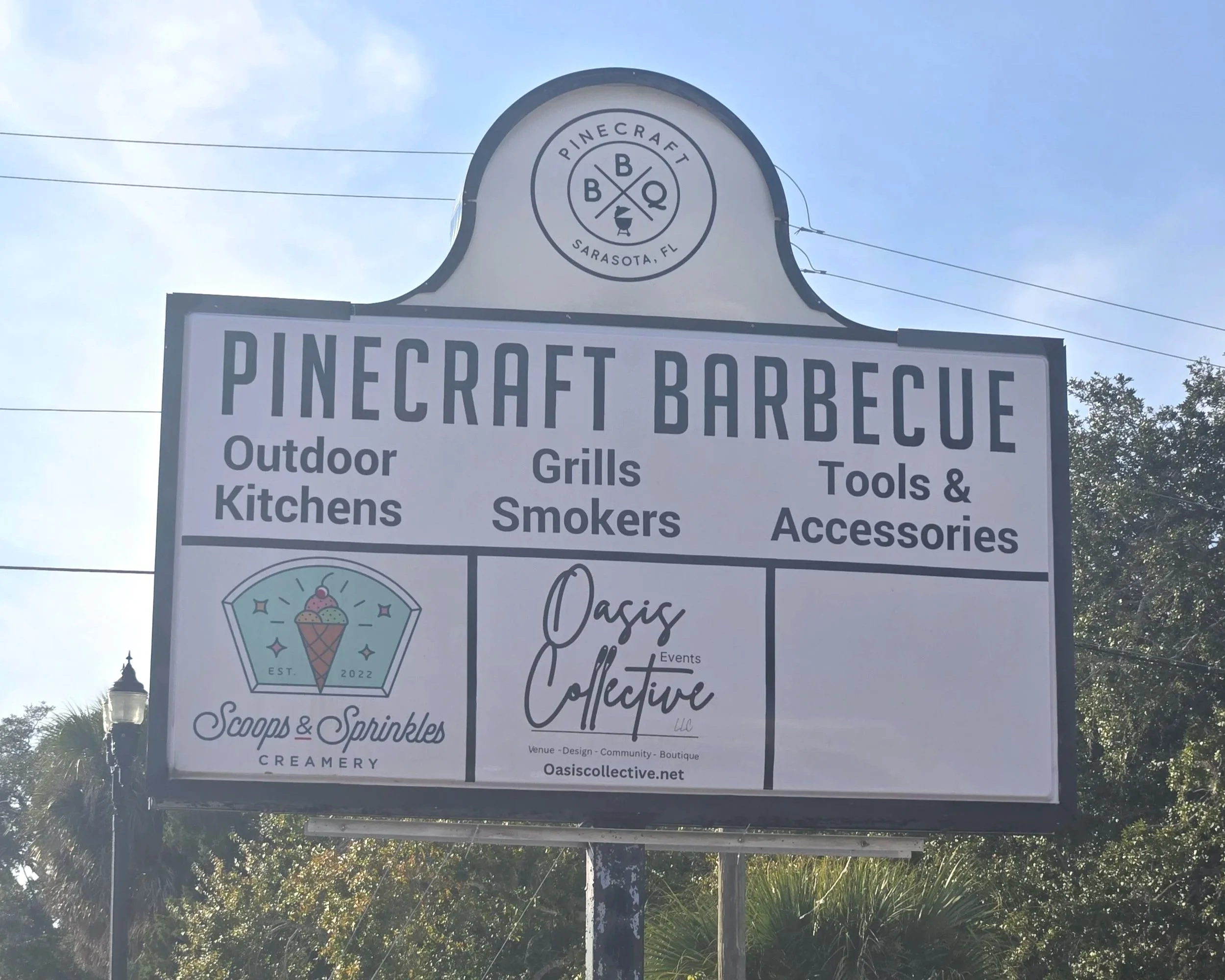 A large outdoor sign for Pinecraft Barbecue in Sarasota, Florida, advertising outdoor kitchens, grills, smokers, tools, and accessories, with additional logos for Scoop & Sprinkles Creamery and Oasis Collective events.