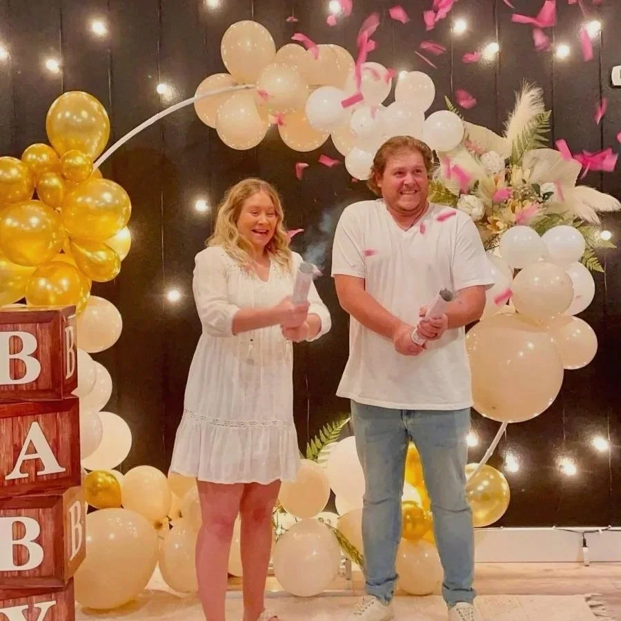 A woman and a man celebrating, standing in front of a decorative backdrop with white and gold balloons, pink confetti, and floral arrangements. The woman is holding a microphone and smiling, while the man is also holding a microphone and smiling.