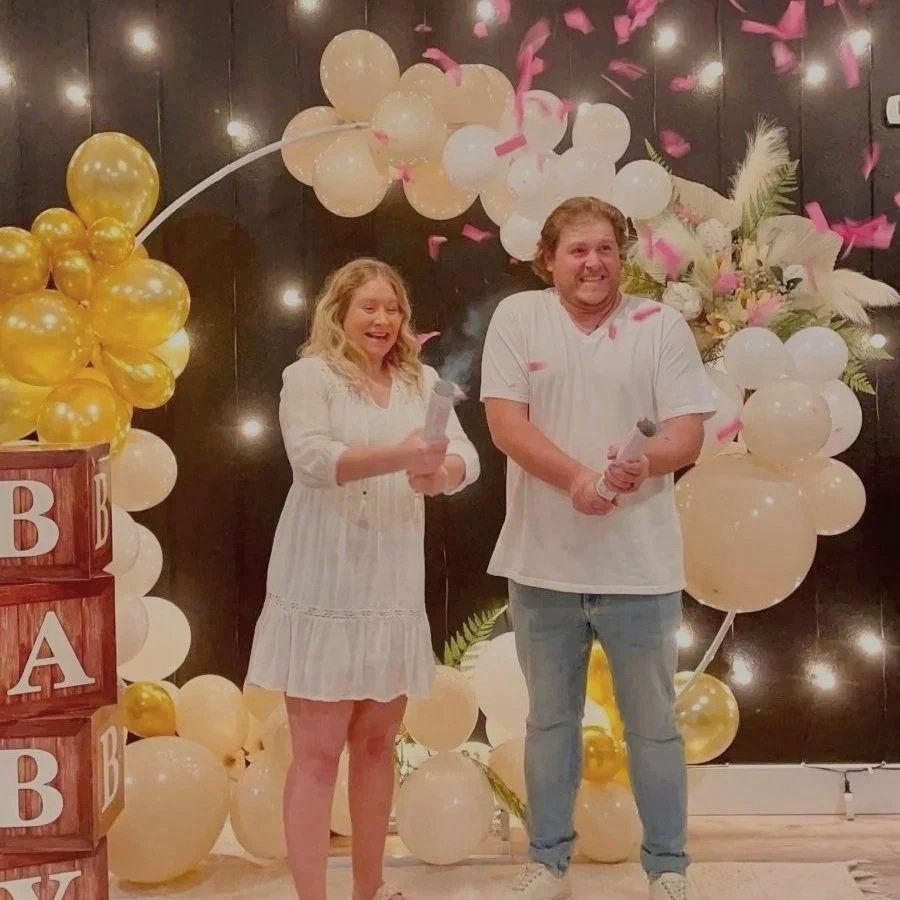 A woman and a man celebrating, standing in front of a decorative backdrop with white and gold balloons, pink confetti, and floral arrangements. The woman is holding a microphone and smiling, while the man is also holding a microphone and smiling.