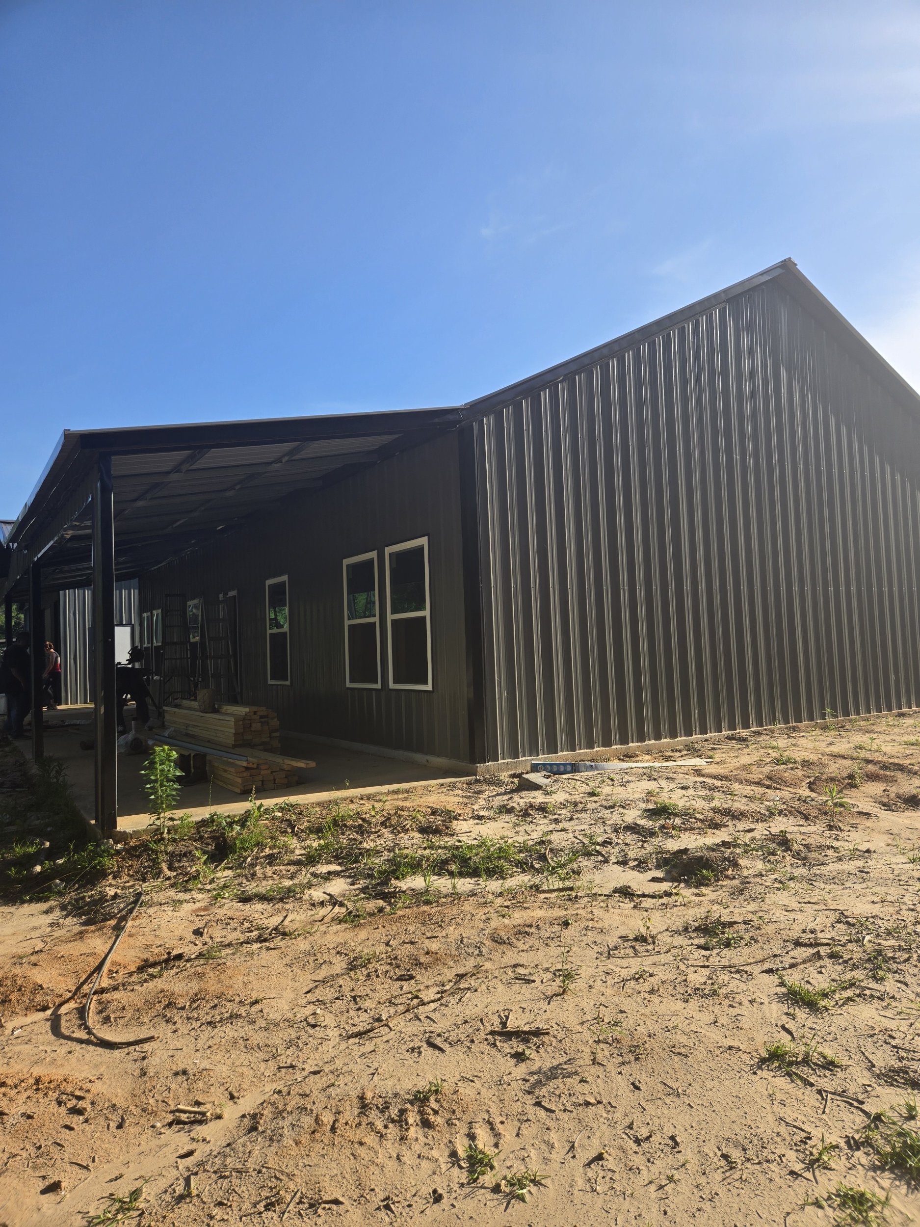 A black metal building under construction with a sloped roof, three windows, and an outdoor covered porch, on a dirt lot with small plants under a clear blue sky.