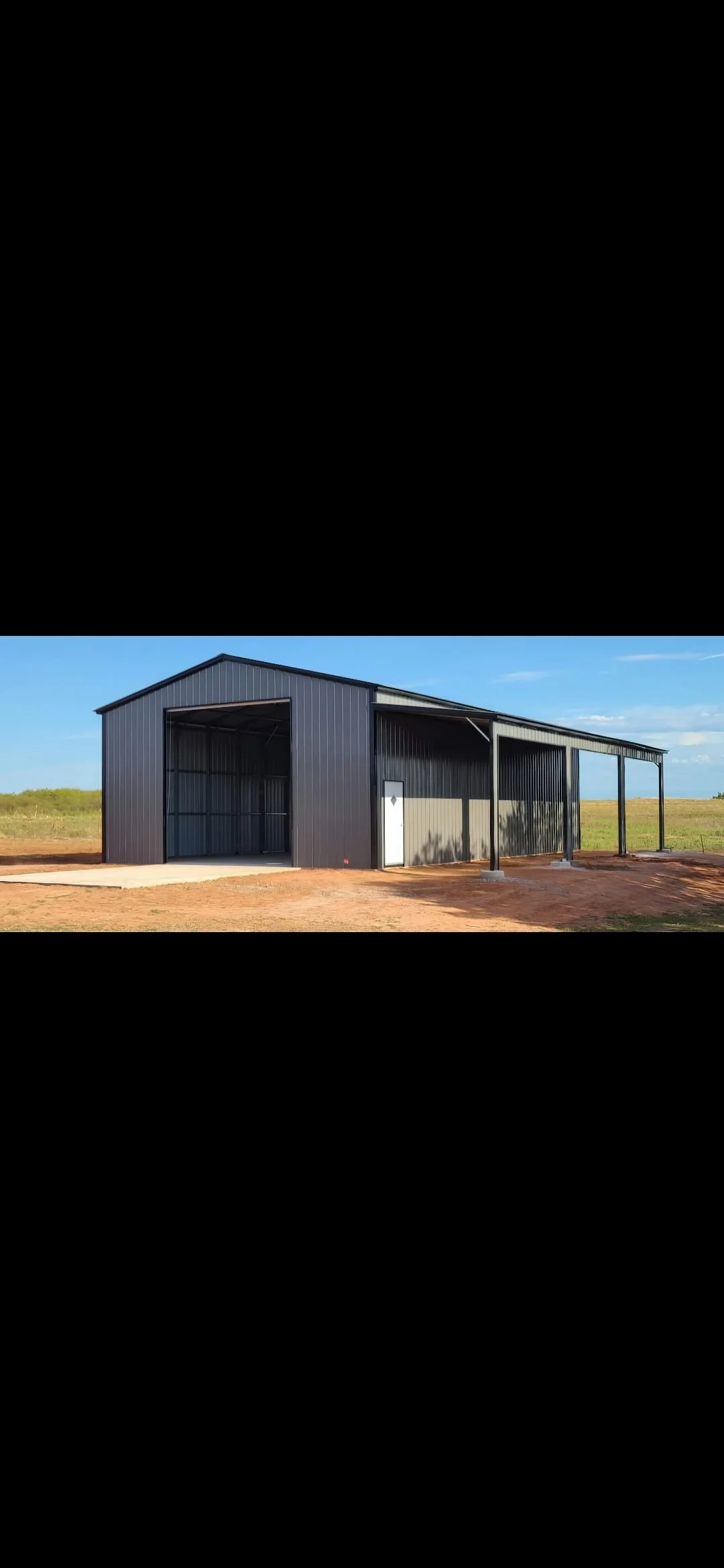 A large, black metal building with an open side and a smaller white door on the side, situated in a rural area with flat land and blue sky in the background.