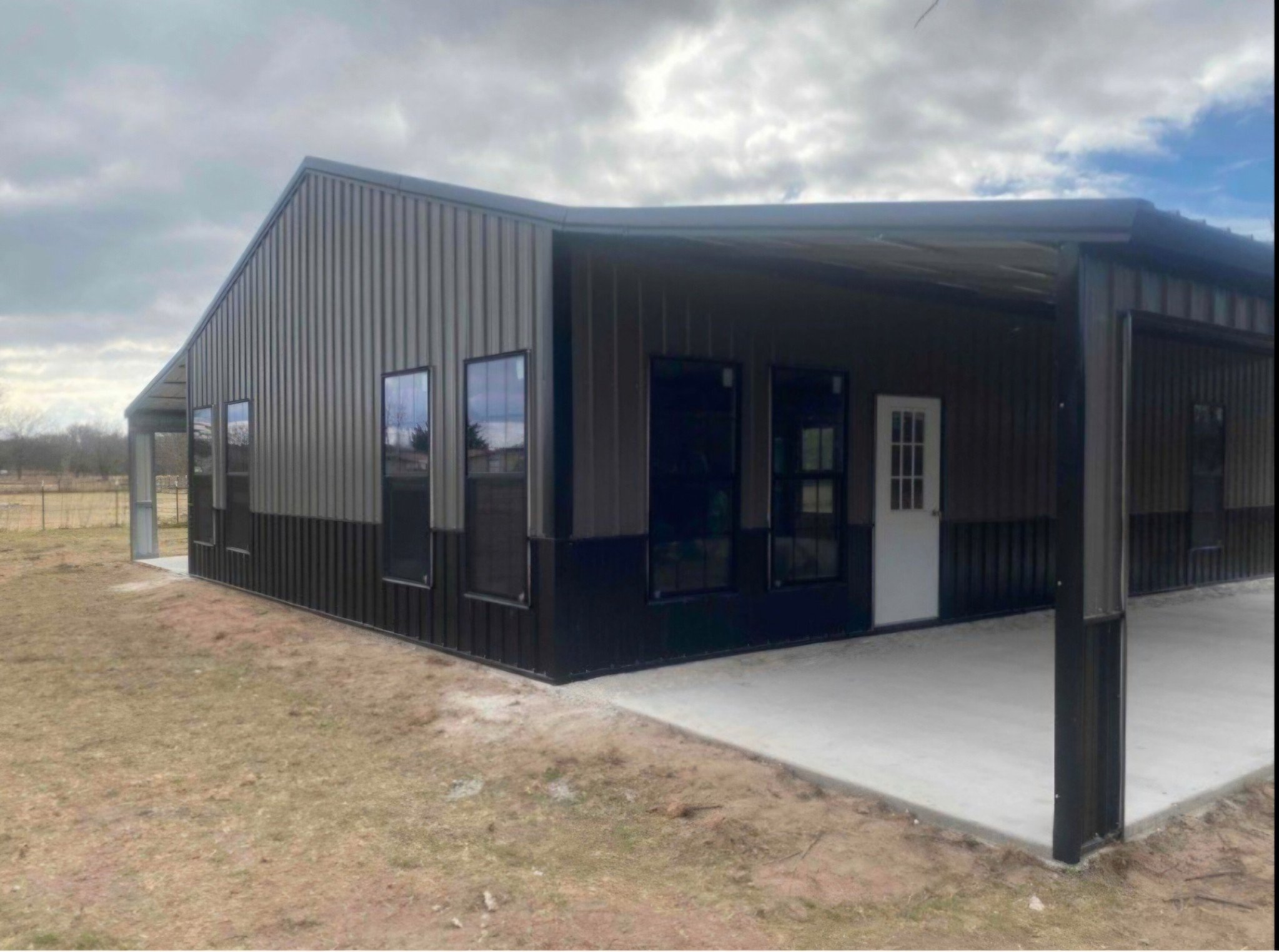 A modern metal building with vertical siding, multiple windows, and a white door, situated on a concrete slab in an open area with some grass and a cloudy sky.