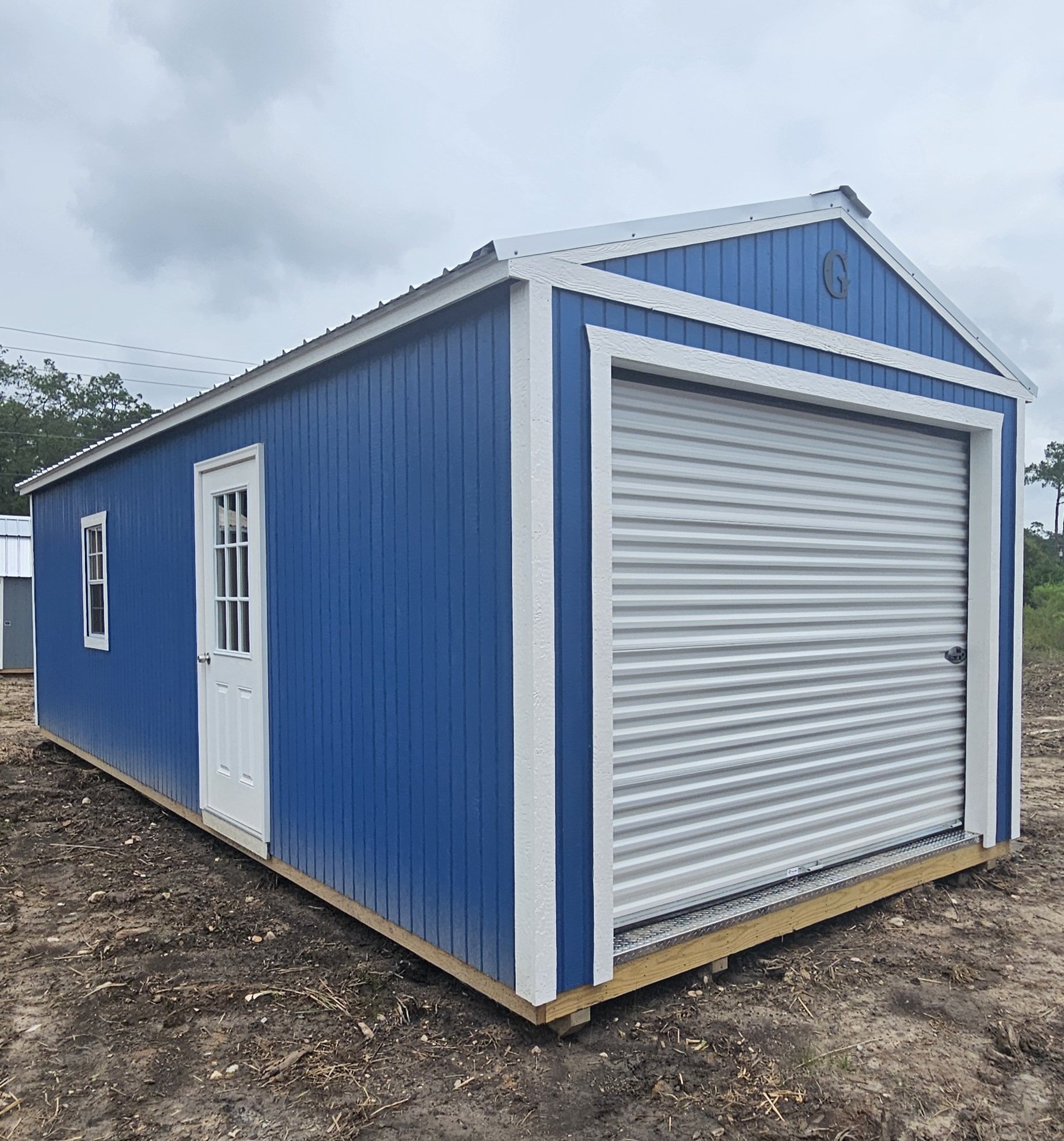 Blue and white metal storage building with a roll-up door and small side door, situated on a wooden foundation in an outdoor area.