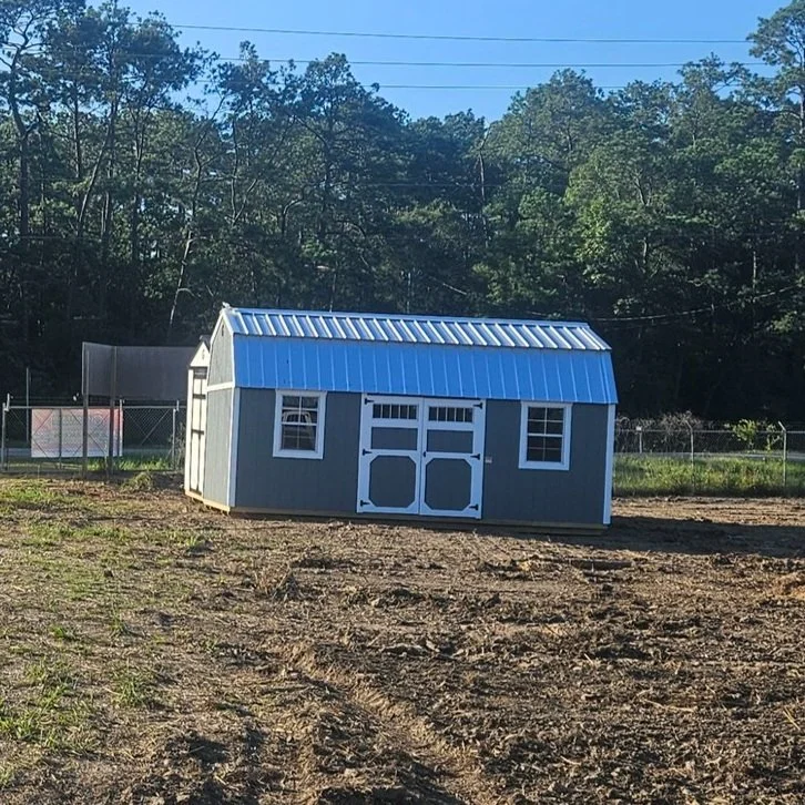 A small gray garden shed with a blue metal roof and white trim, situated in a dirt yard with a fence and trees in the background.