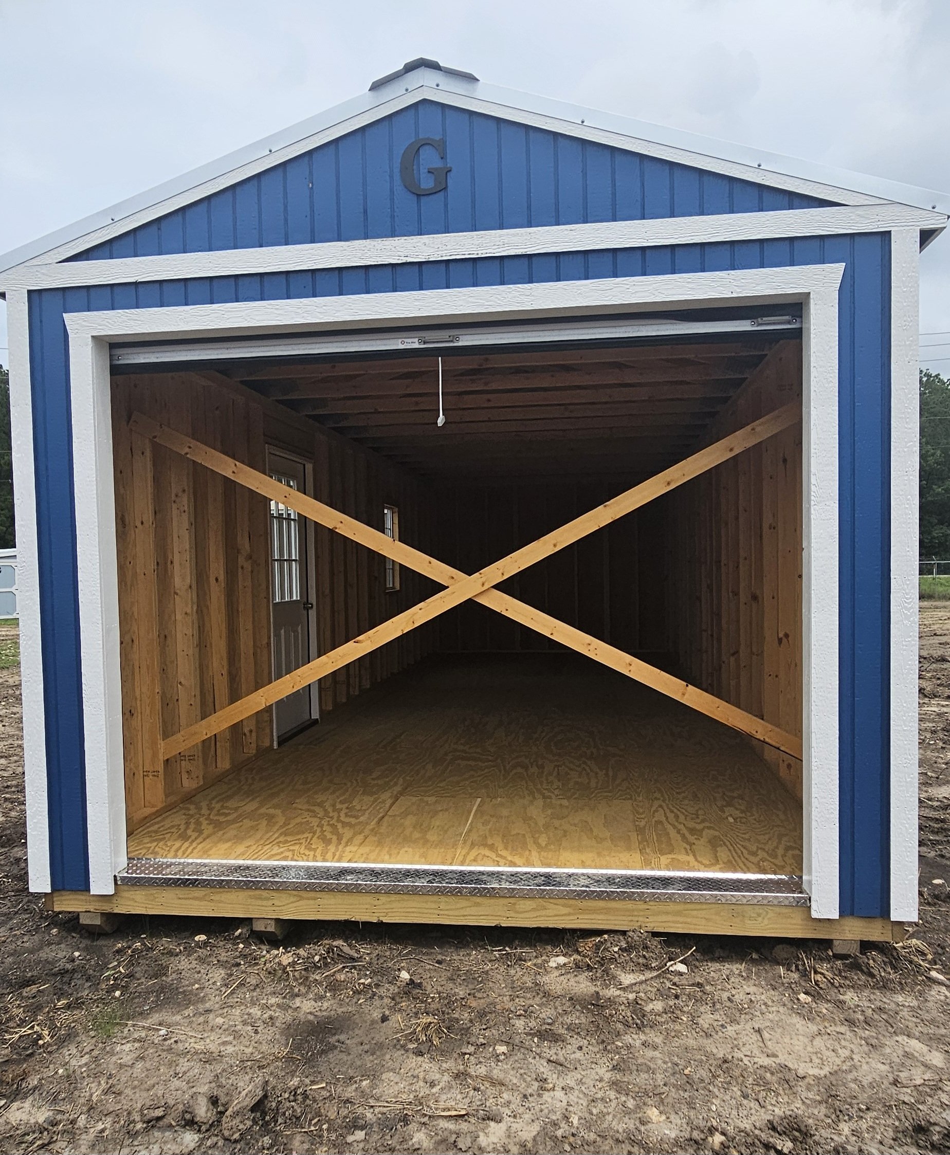 Empty blue and white storage shed with wooden interior and two small windows, with a cross brace on the front opening, on a patch of dirt ground.