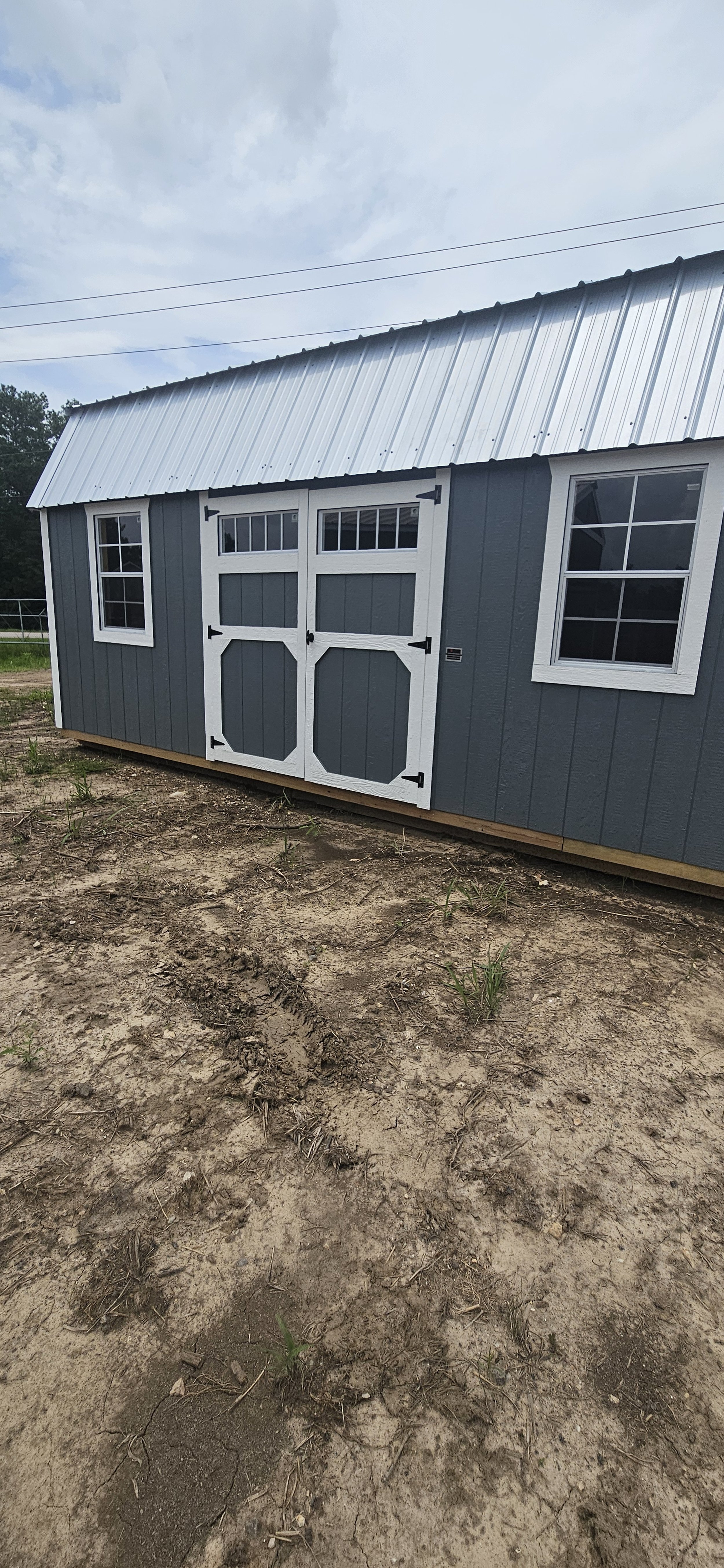 A gray shed with white trim and black hardware, featuring two windows and a pair of barn-style doors, situated on dirt ground under a cloudy sky.