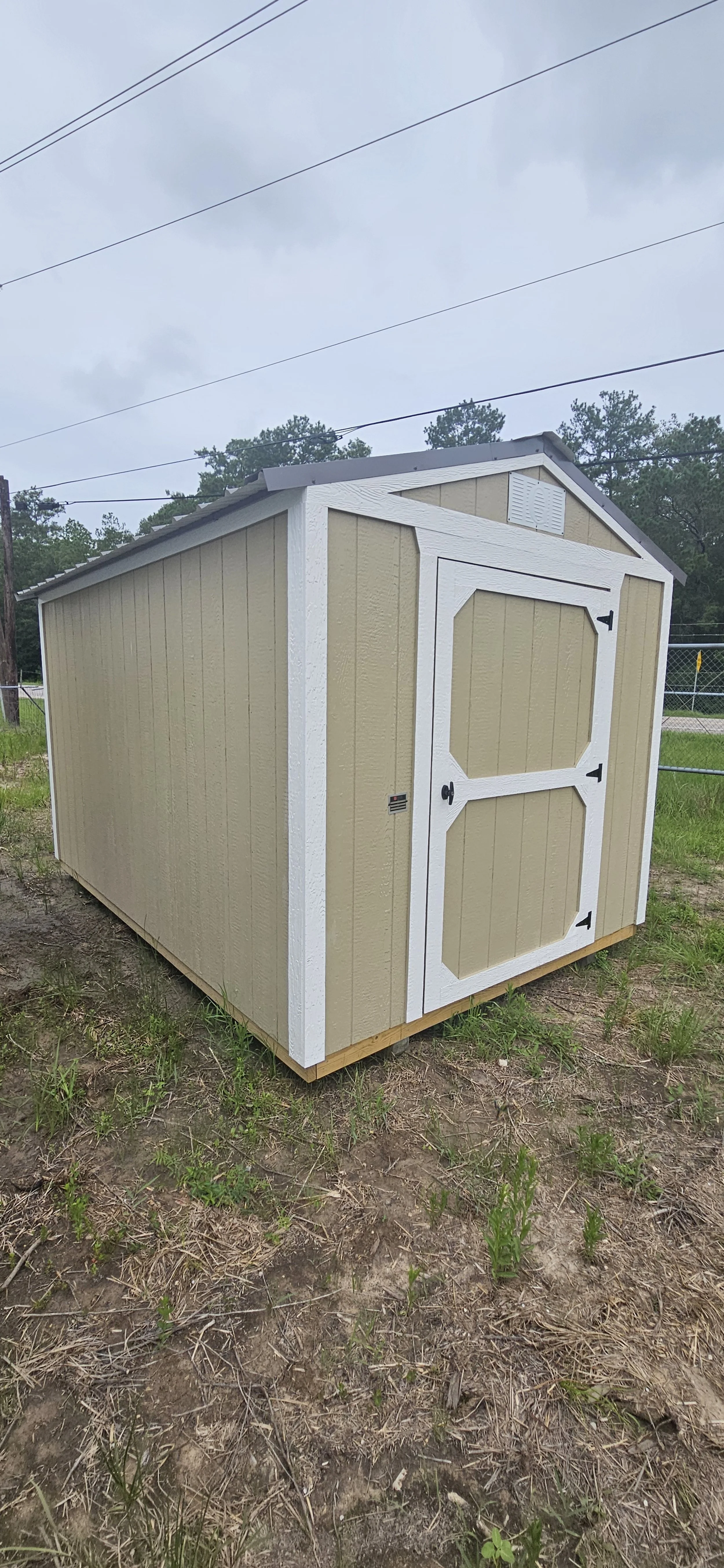 A small beige outdoor storage shed with white trim and a double door, situated on a patch of dirt and grass.