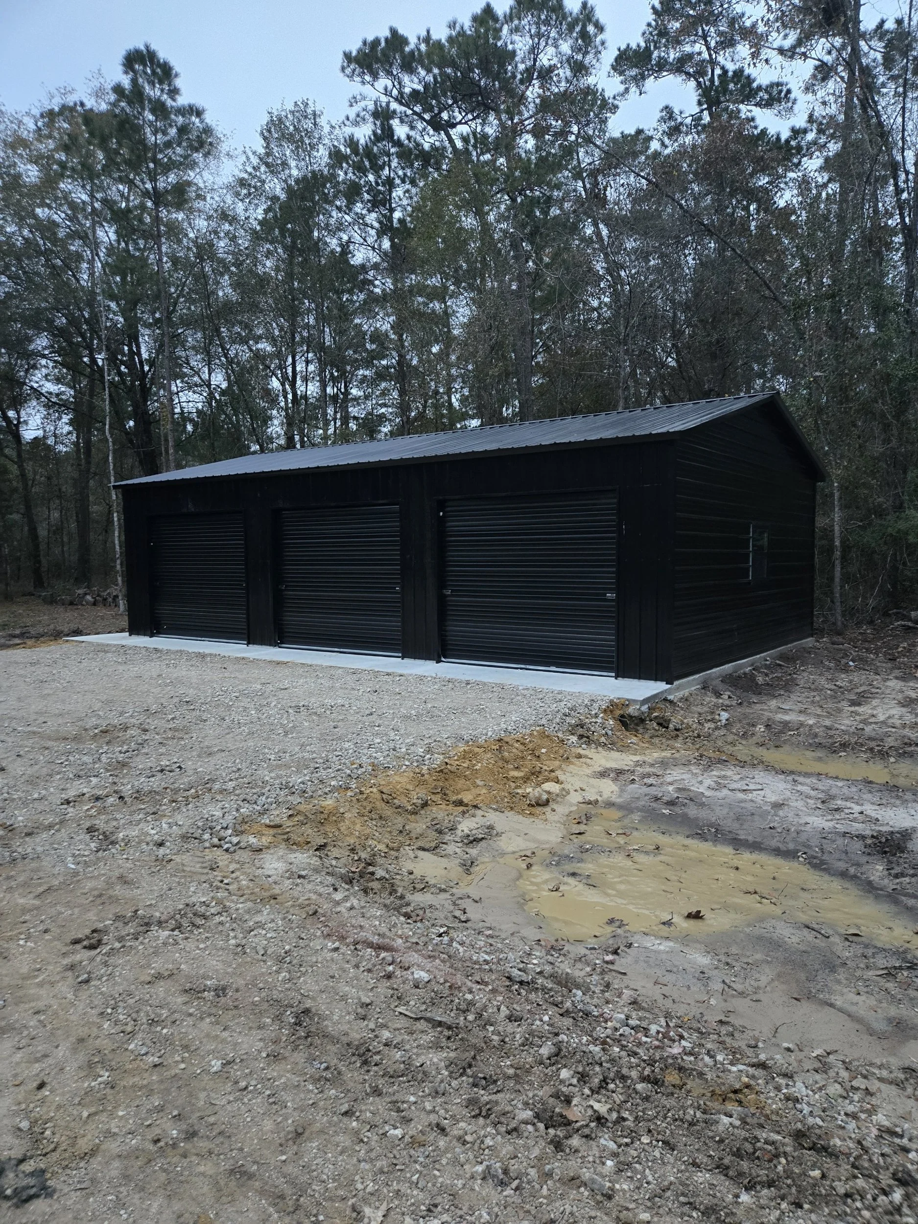 A black metal storage building with three roll-up garage doors, situated on a gravel foundation, in a wooded area with a dirt ground in the foreground.