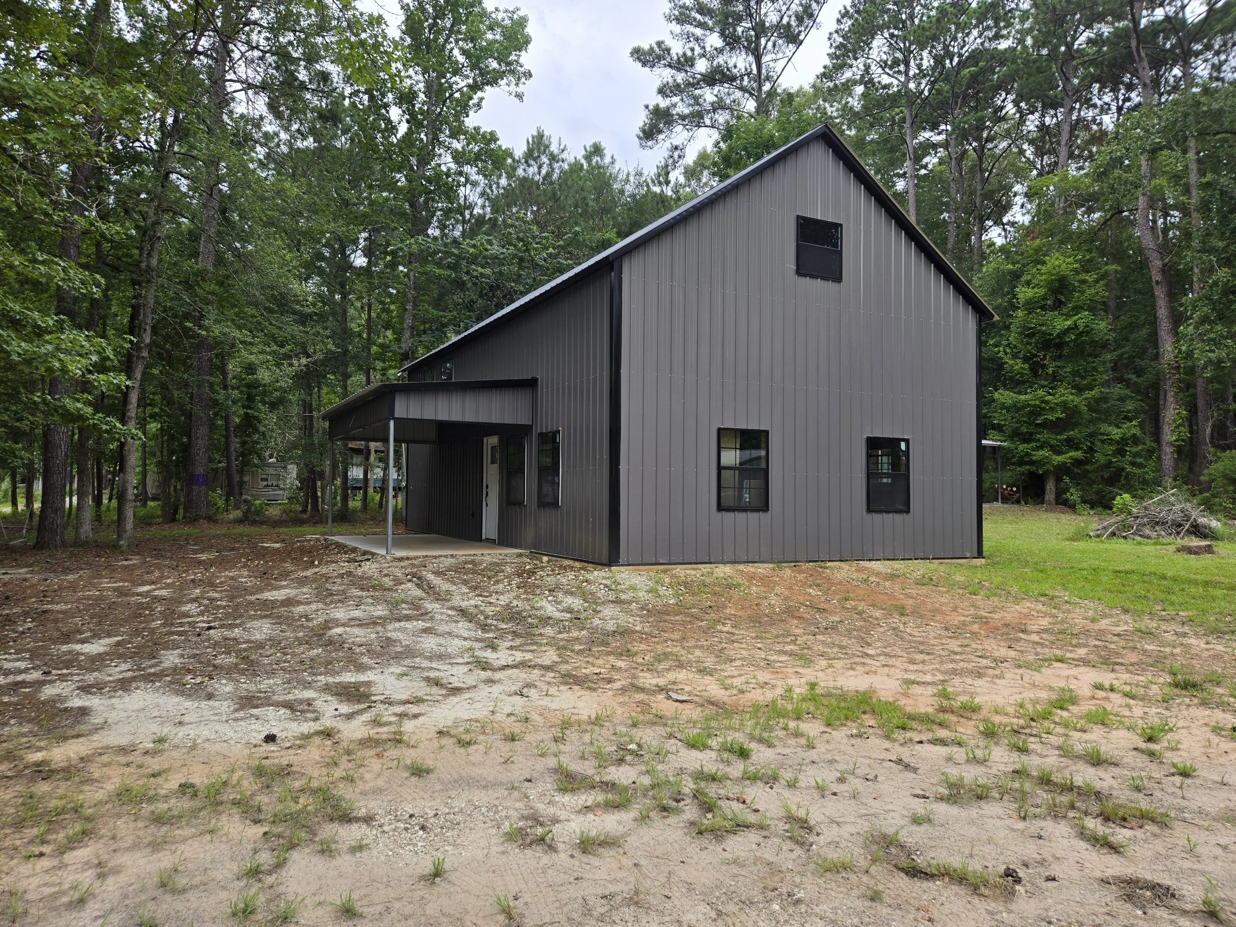 A modern gray metal house with a sloped roof, surrounded by trees, with a small covered porch area and multiple black-framed windows.