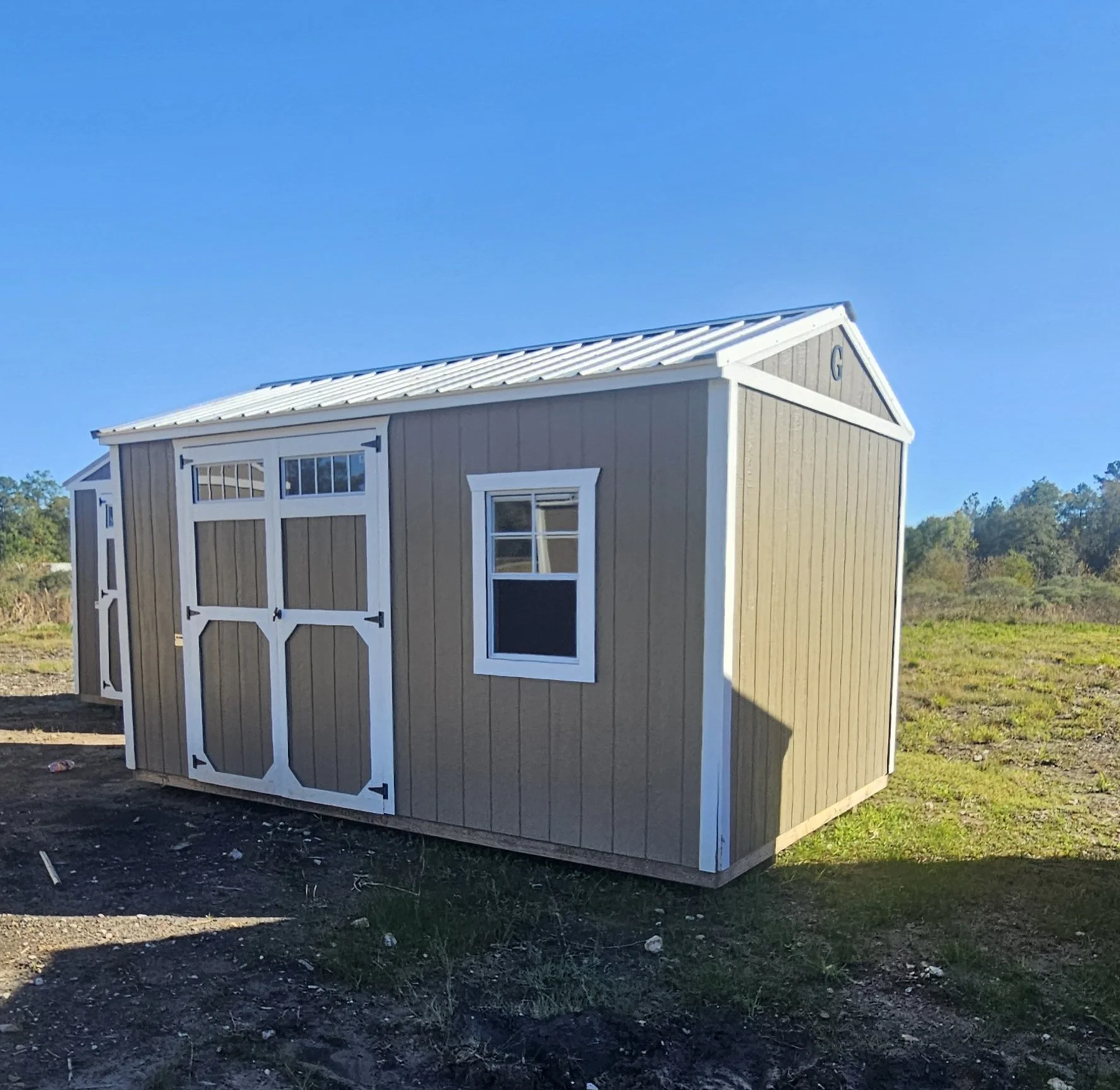 A tan and white shed with white trim, a metal roof, a window, and a sliding barn-style door, situated on a grassy and dirt area under a clear blue sky.