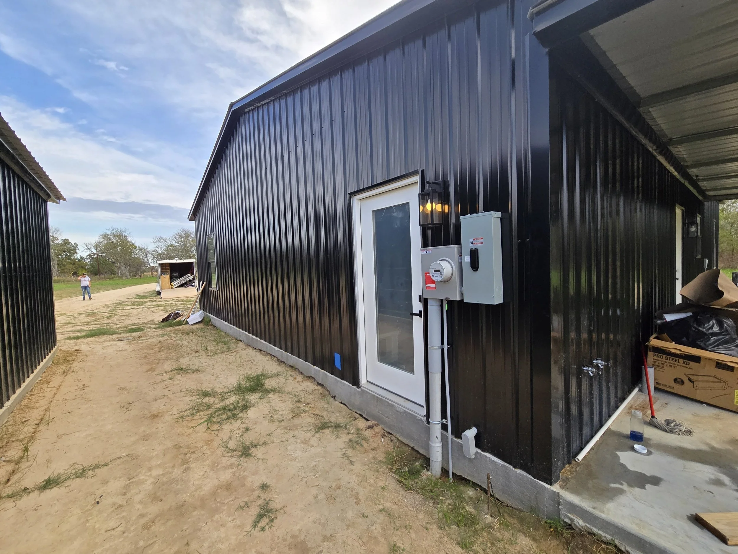 Exterior view of a black metal building with a white door, electrical meters, and an outdoor light fixture. The area around the building has dirt and sparse grass, with construction materials and tools nearby.
