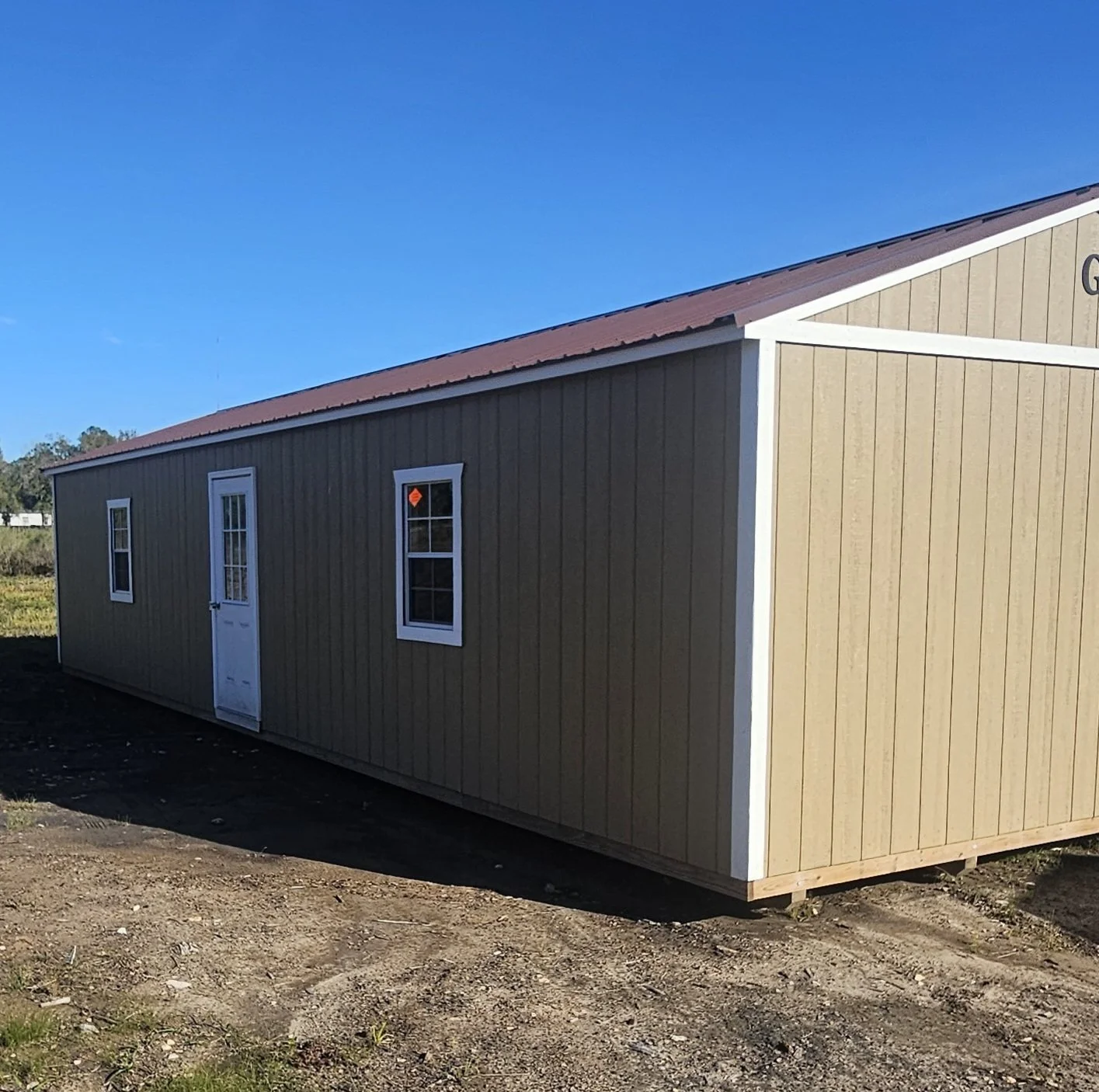 A beige rectangular portable building with white trim, two windows, and a door, set on a dirt lot under a blue sky.