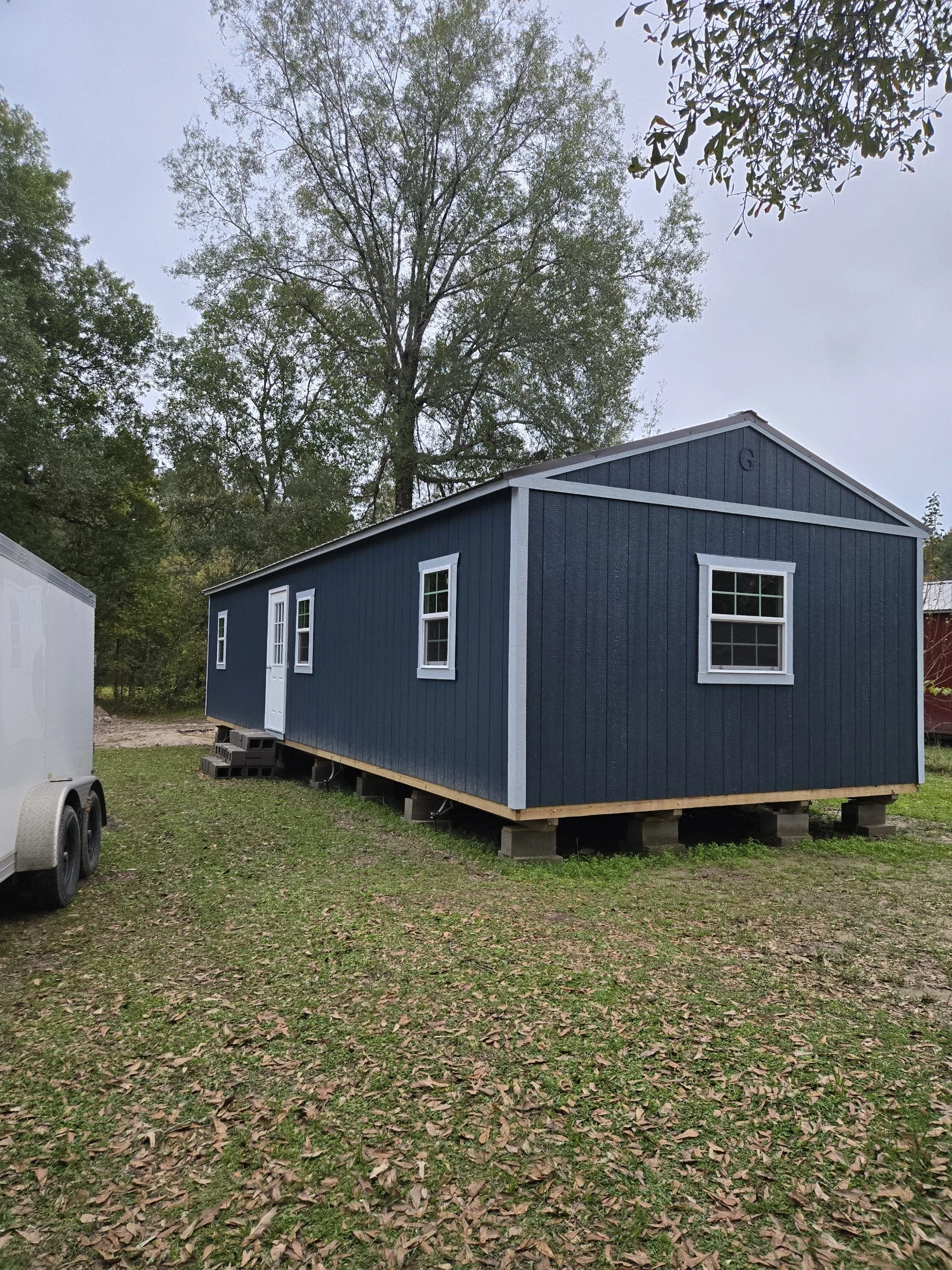 Blue modular house raised on cinder blocks in a grassy area with trees in the background.