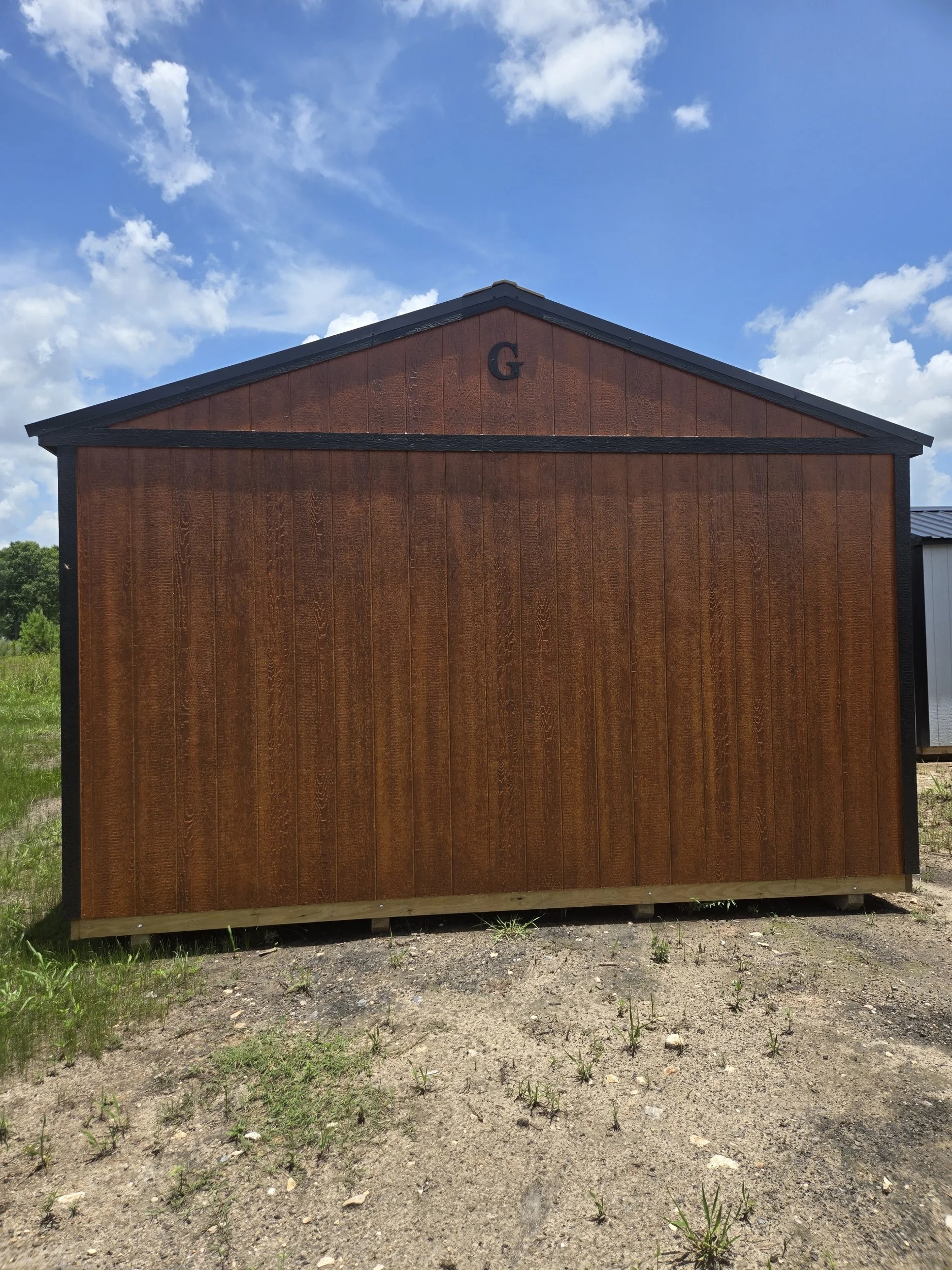 A small brown shed with a black roof and trim, situated outdoors under a partly cloudy sky, with a green field in the background and a lone letter G on the upper front.