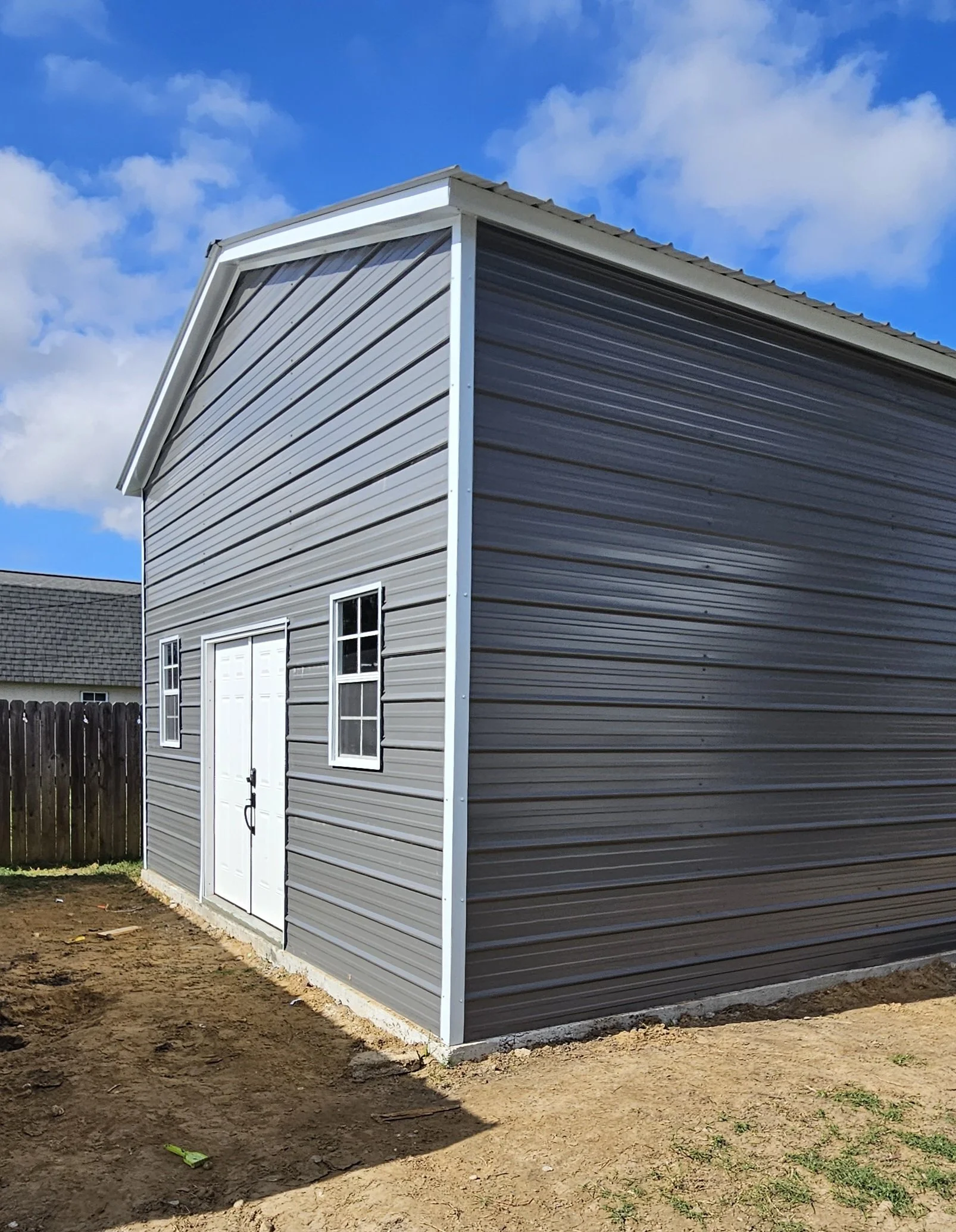 A gray metal shed with white doors and windows, situated on a patch of dirt in a backyard, under a partly cloudy sky.