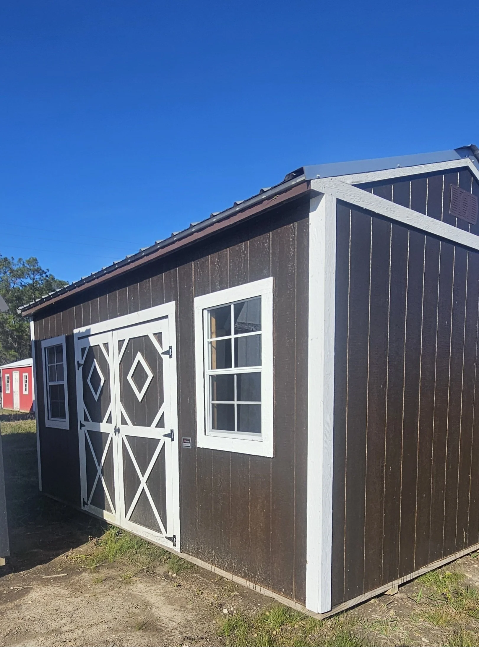 A small brown shed with white trim and double doors featuring decorative designs. The shed has two windows with white frames, set on a grassy and dirt area under a clear blue sky.