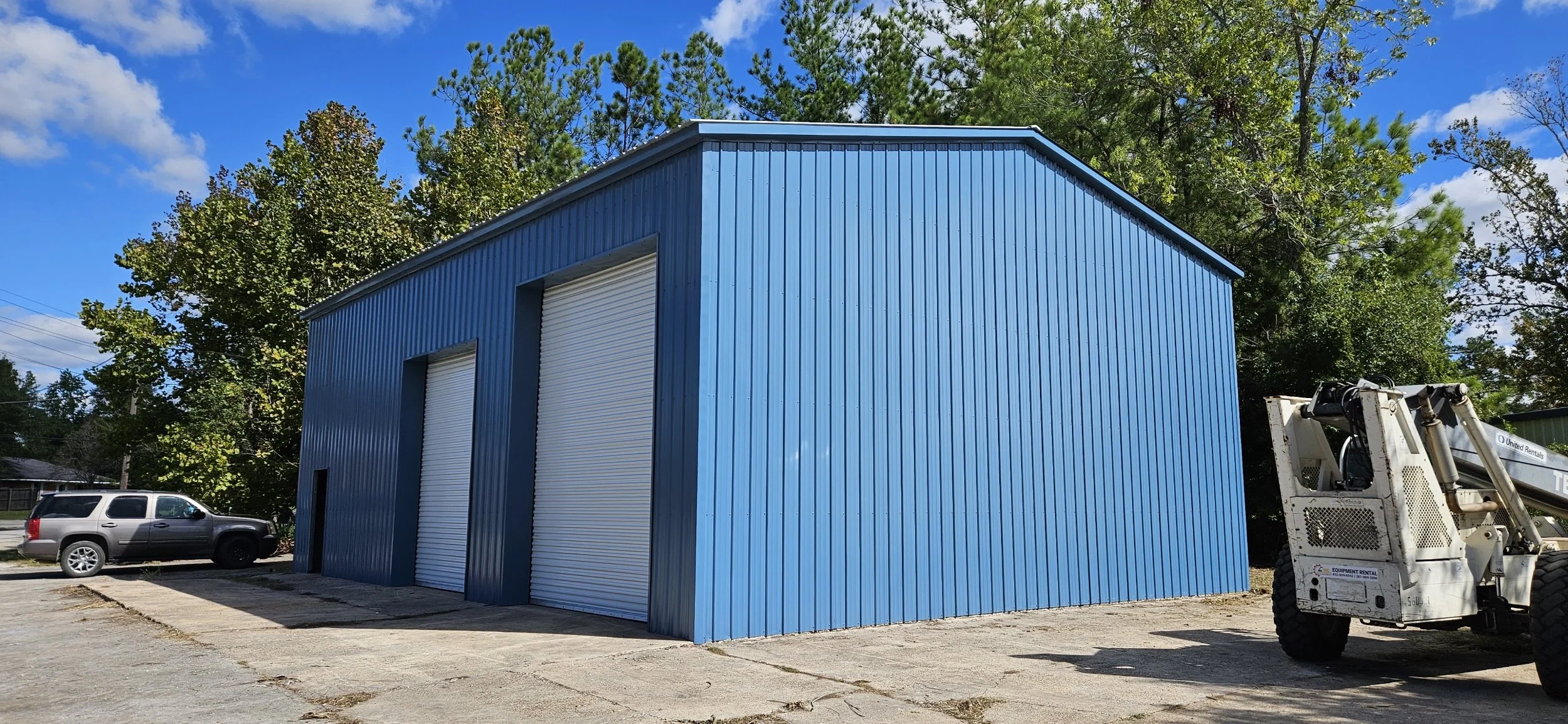 Blue metal storage building with two large roll-up doors, a smaller door, and a white construction vehicle parked nearby, with trees and a blue sky in the background.
