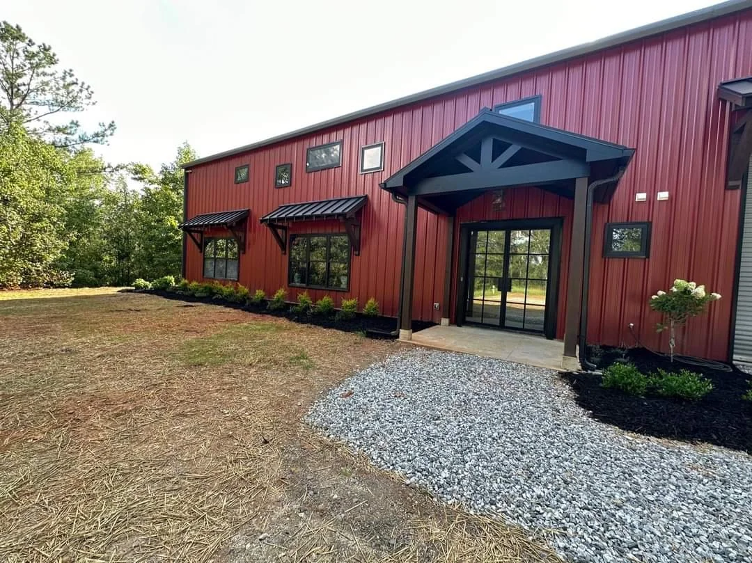 Red barn-style building with black window awnings, a glass door with a small porch, and a gravel pathway leading to it, surrounded by trees and new landscaping.