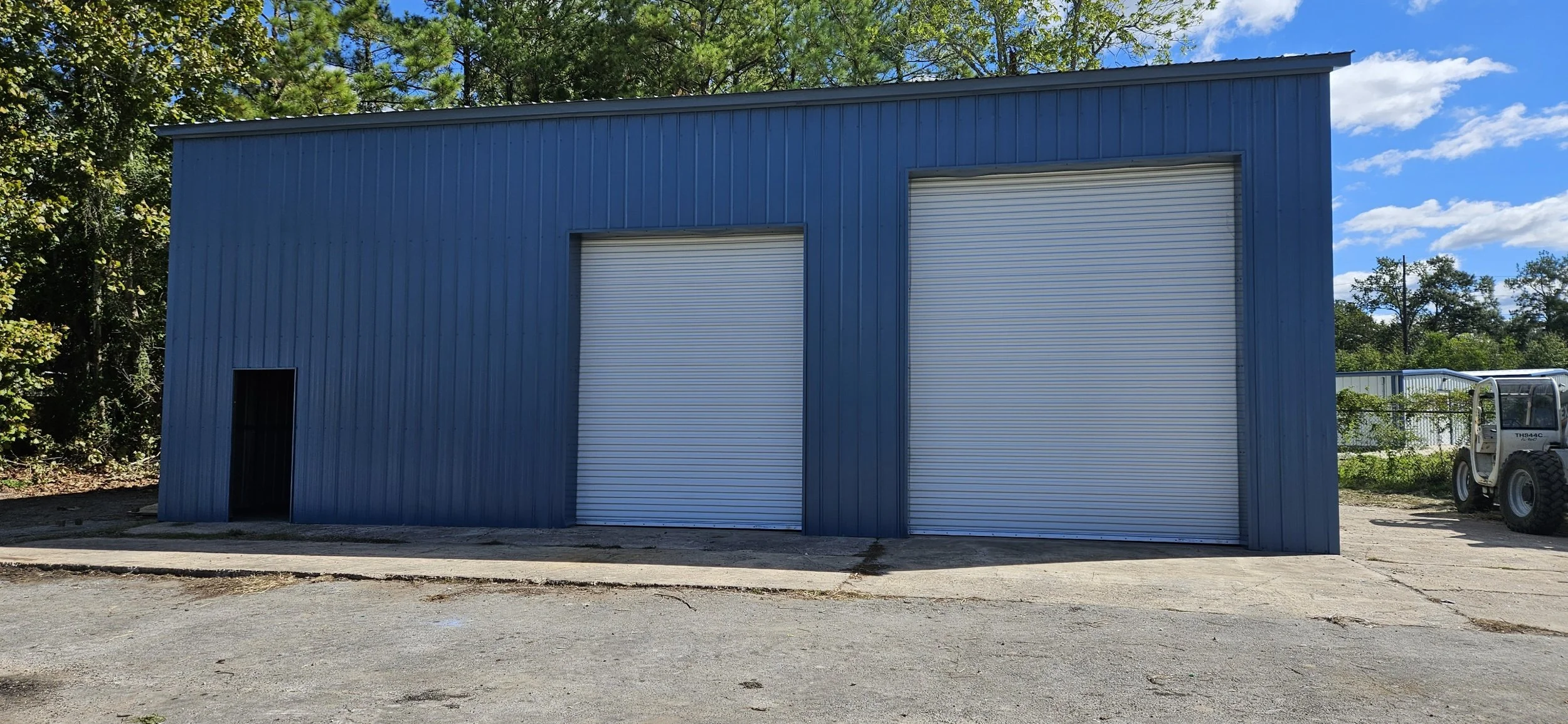 Blue metal industrial building with two large roll-up doors and a small side door, situated on a concrete lot with trees in the background and a partly cloudy sky above.