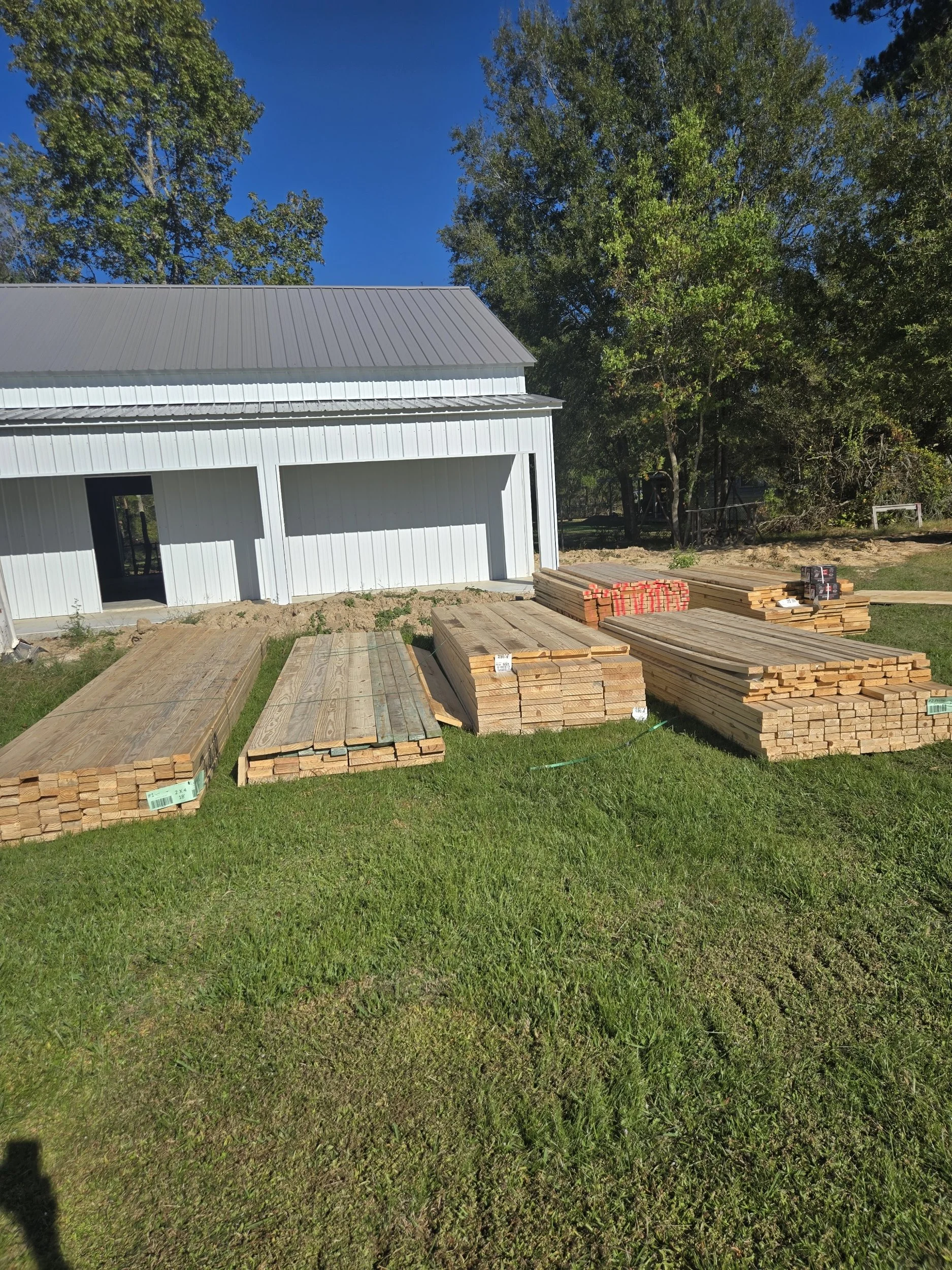 Stacks of wooden planks and lumber on a grassy area in front of a white metal building with a gray roof, surrounded by trees.