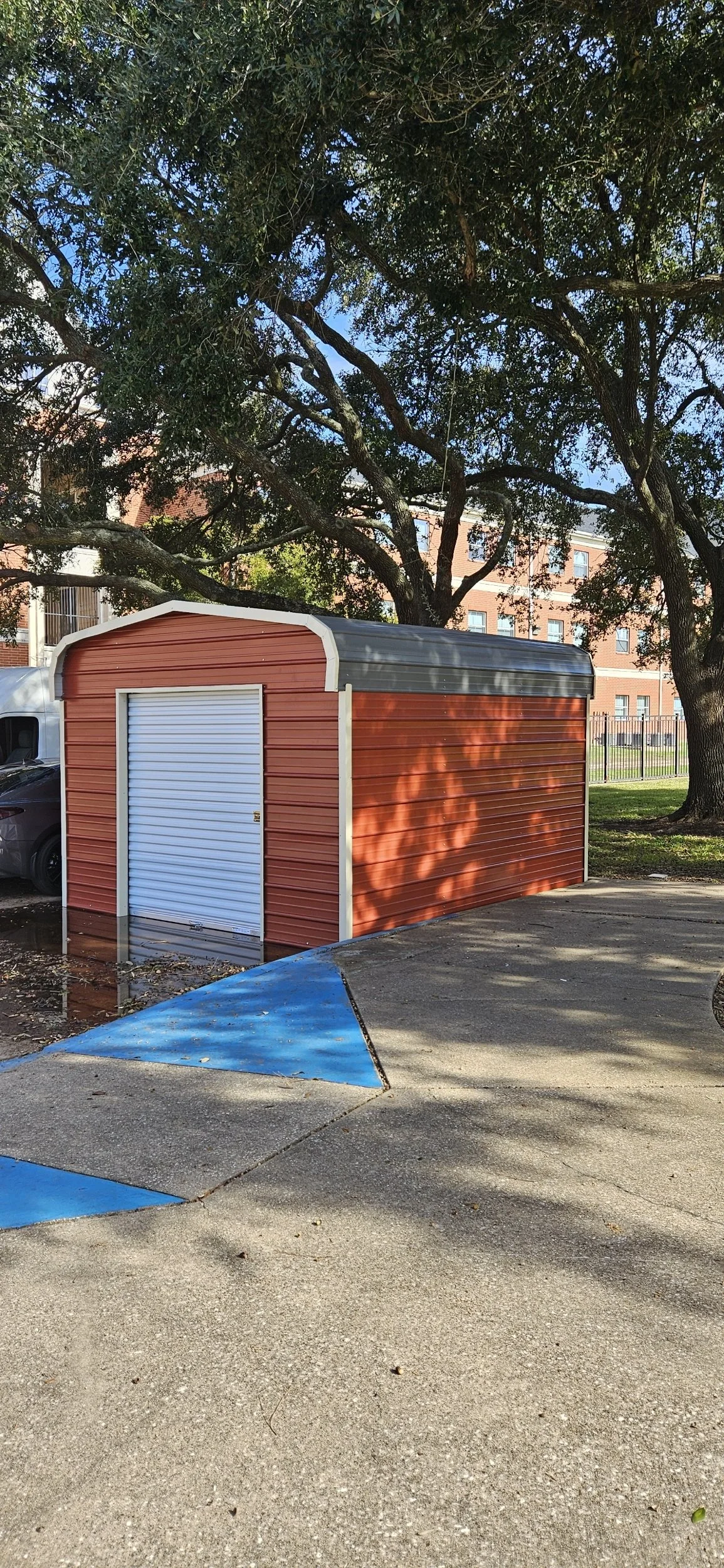 Red metal storage shed with a white roll-up door, situated on a concrete parking lot with blue-painted parking space lines, under large leafy trees and next to a multi-story building.