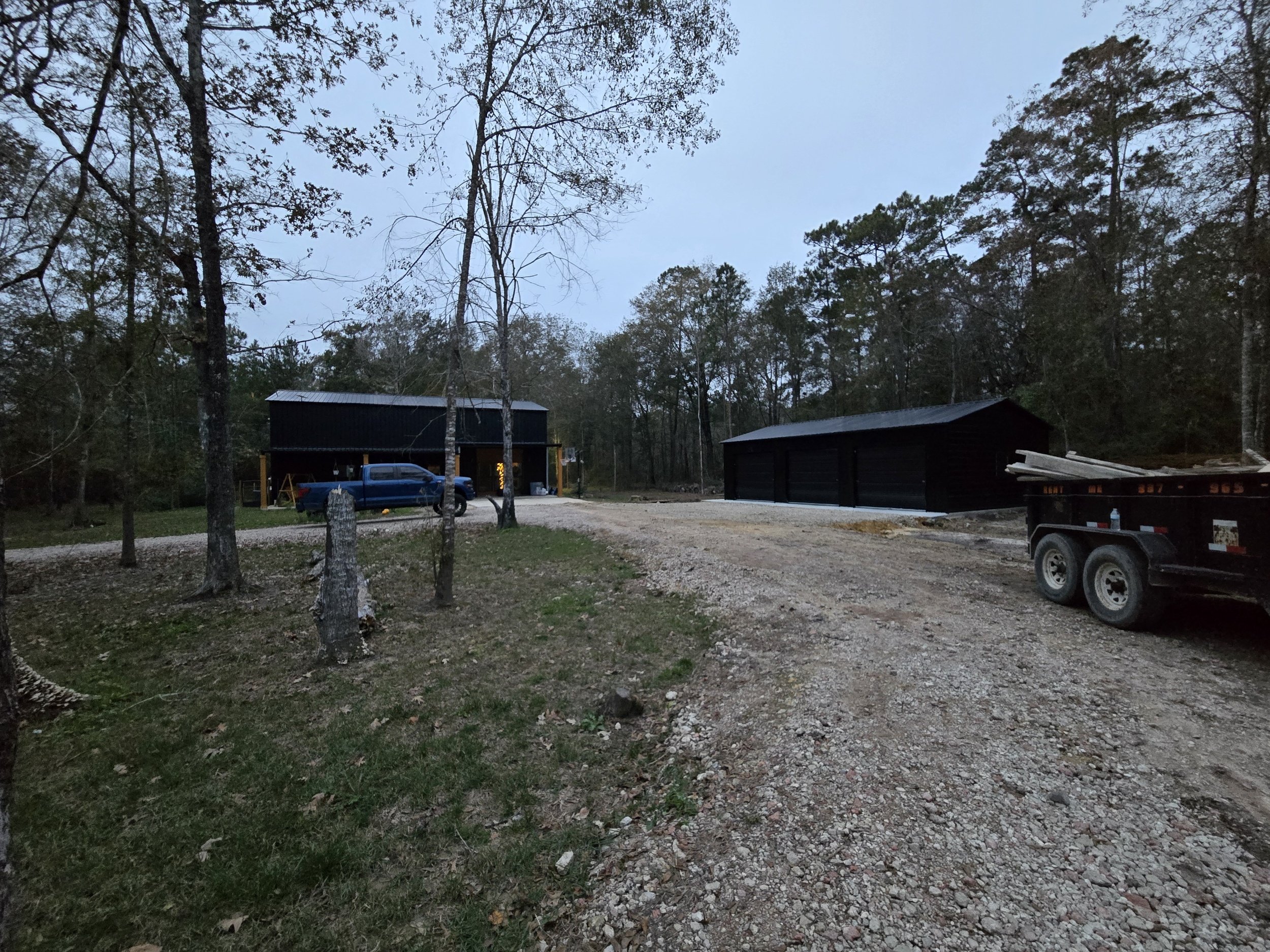 A rural scene during dusk with two black sheds, a blue truck parked near one shed, and a trailer loaded with white PVC pipes on a gravel driveway surrounded by trees.