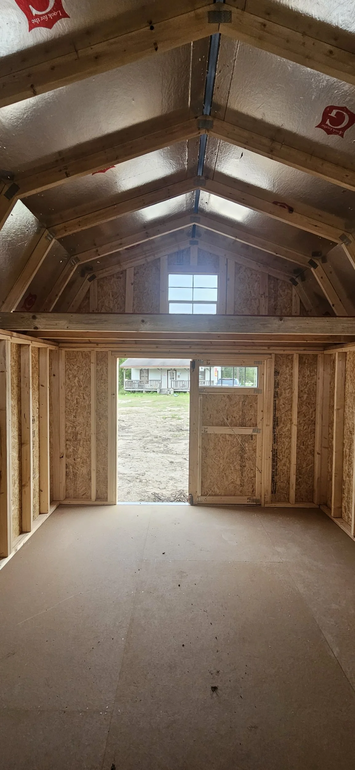 Interior of a small wooden building under construction, showing unfinished walls with framing and insulation, a doorway opening to the outside, and a window on the rear wall, with an elevated loft space.