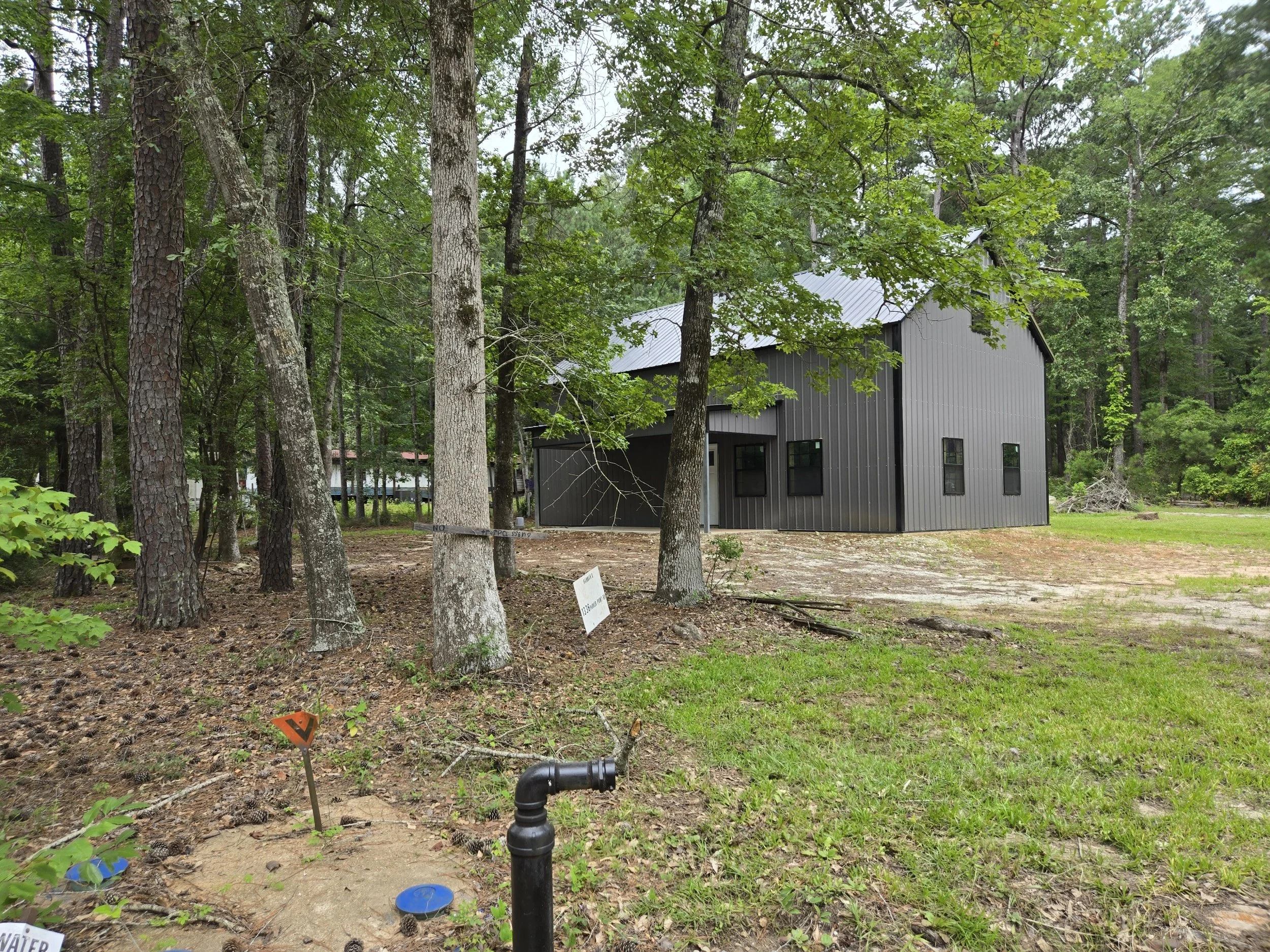 A black barn-style building surrounded by trees and greenery, with a gravel area in front, and some signs and pipes visible in the foreground.