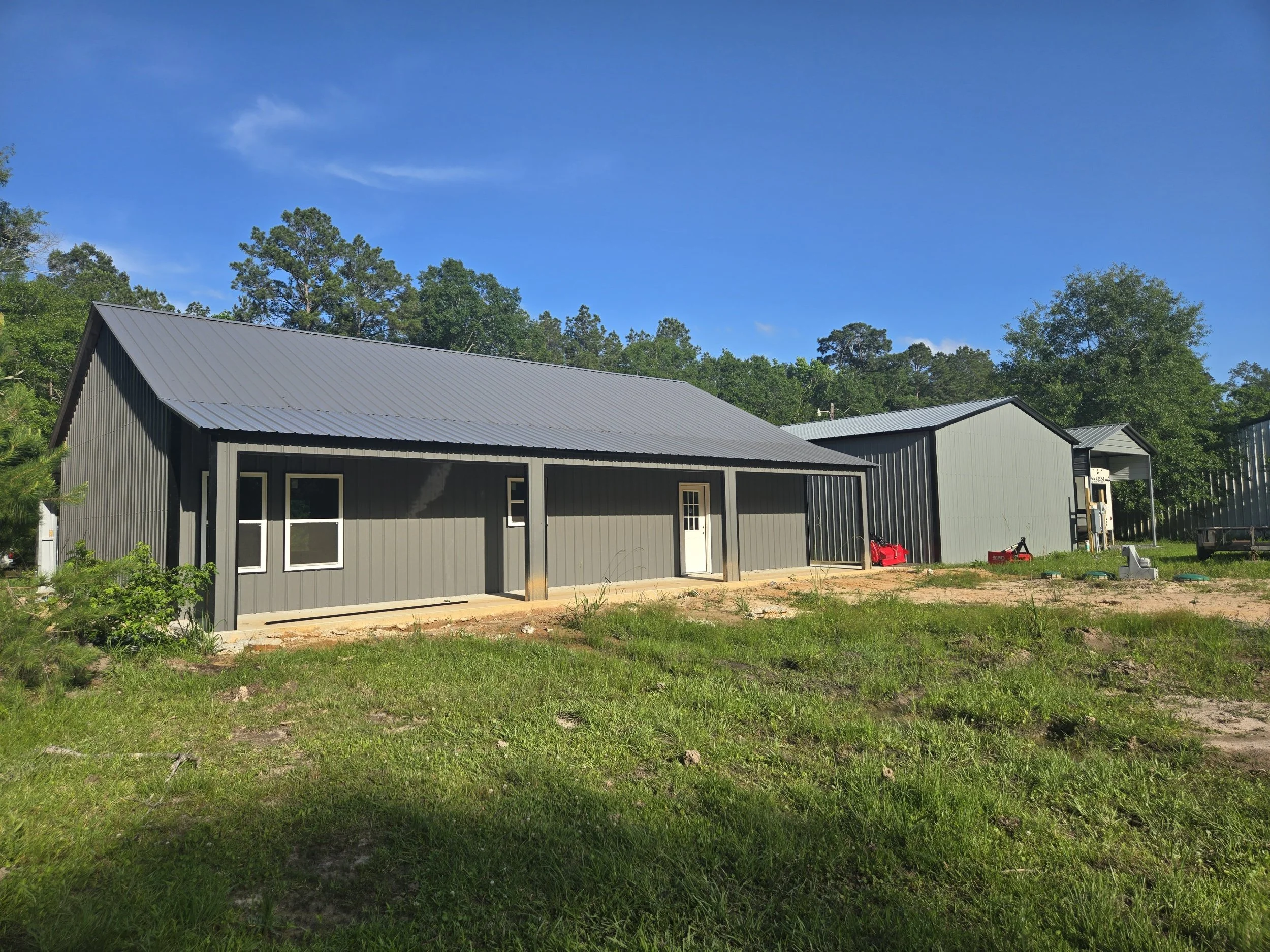 New gray metal buildings with sloped roofs on a grassy lot with trees in the background under a blue sky.