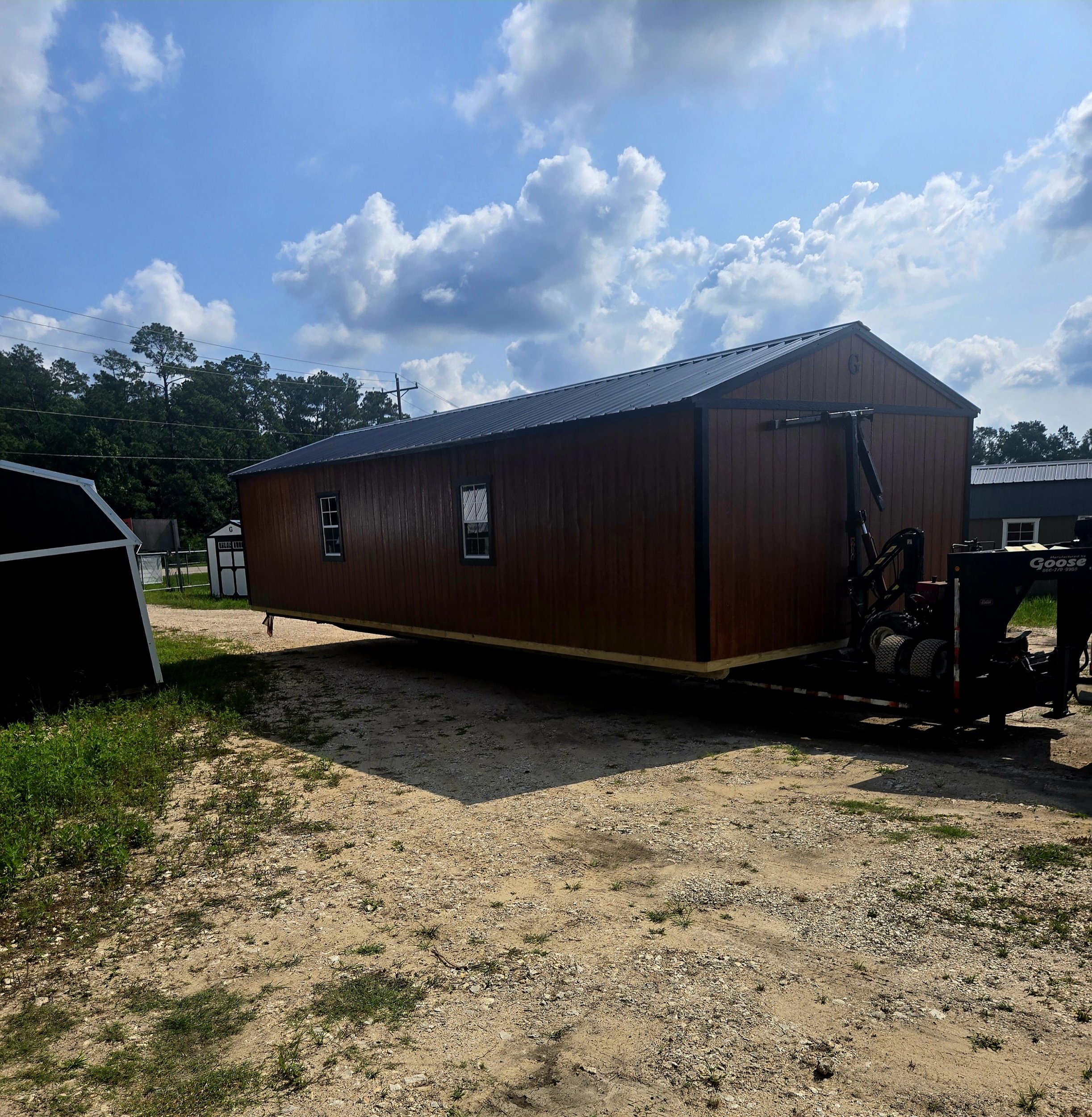 A brown tiny house on wheels with a metal roof, parked on a dirt lot under a partly cloudy sky.