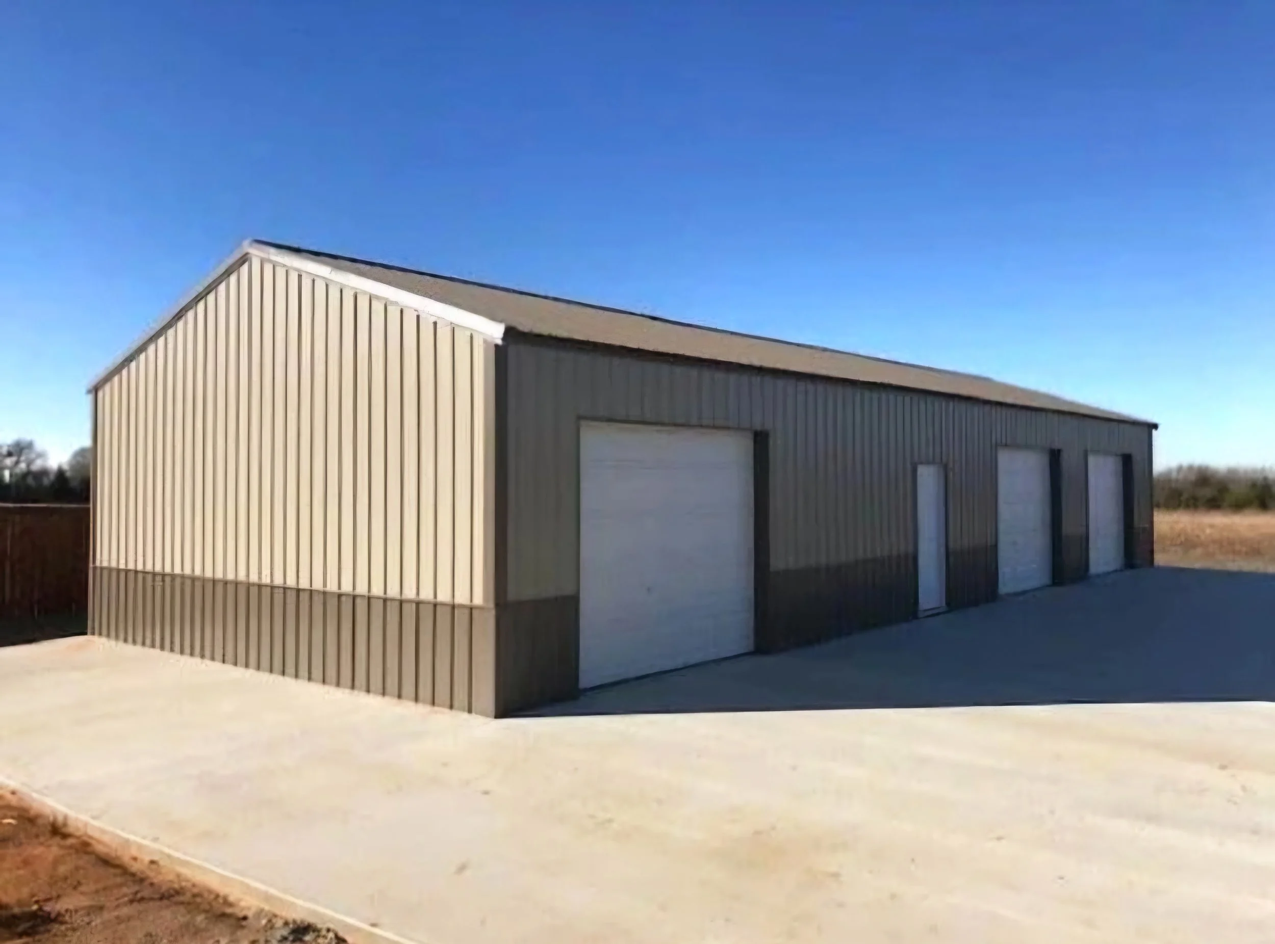 A large metal storage building with three white garage doors, beige upper walls, and darker lower walls, situated on a concrete slab under a clear blue sky.