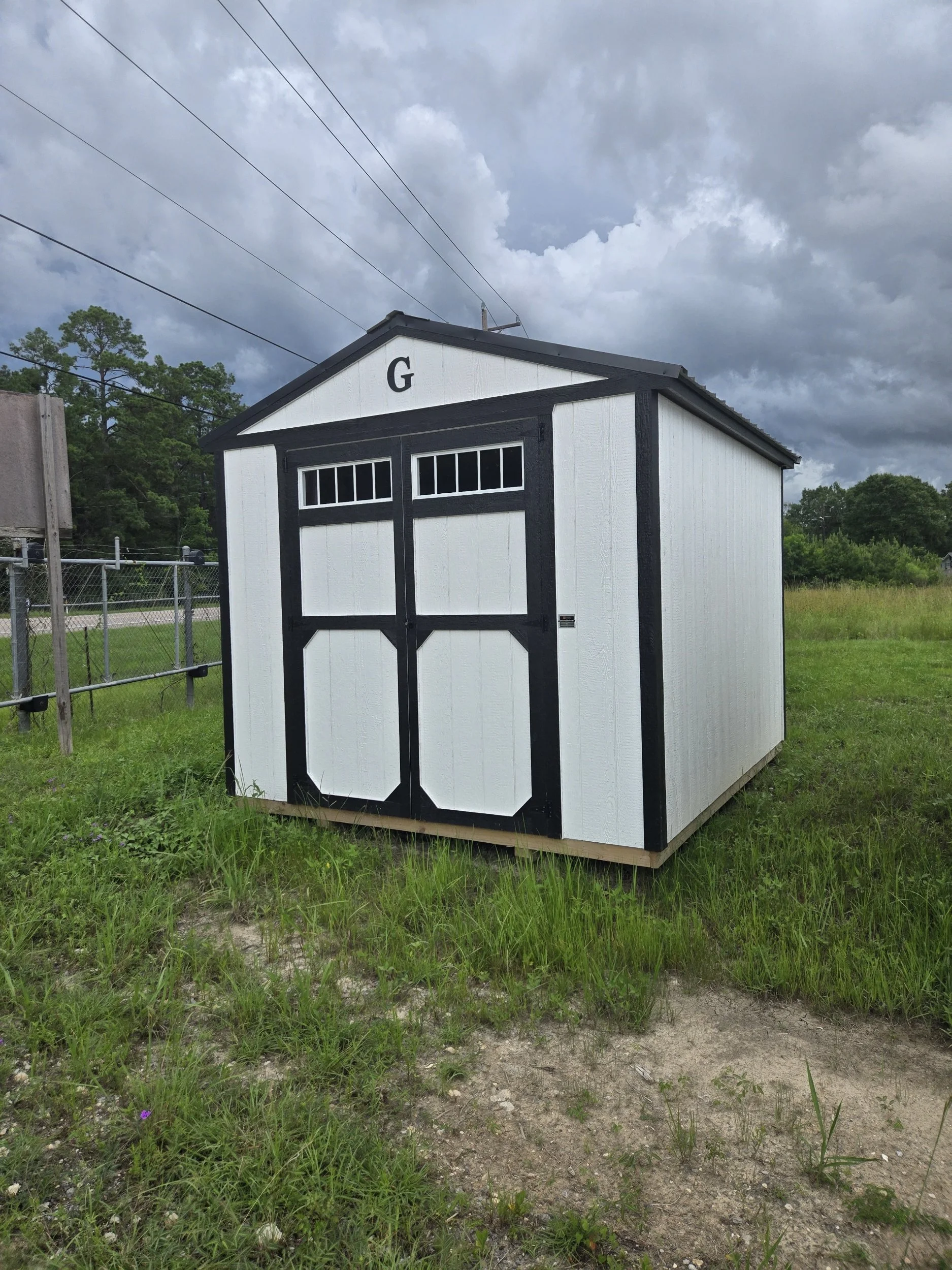 A small white storage shed with black trim and a split double door, situated on grass under a cloudy sky.
