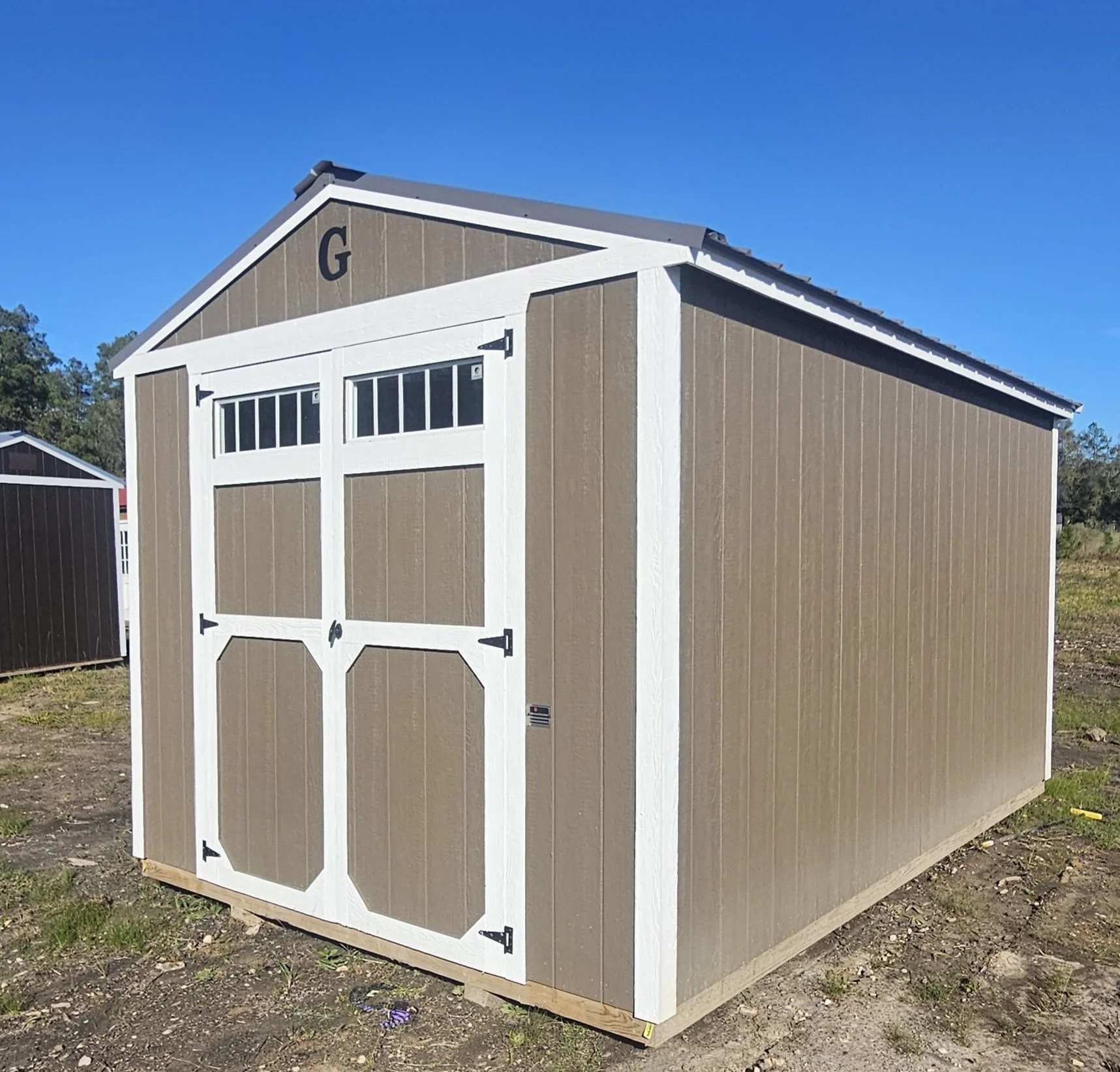 A brown and white shed with a double door, situated on uneven ground in an outdoor area. The shed has a gable roof and small windows at the top of the doors, with a large 'G' letter painted on the front gable.