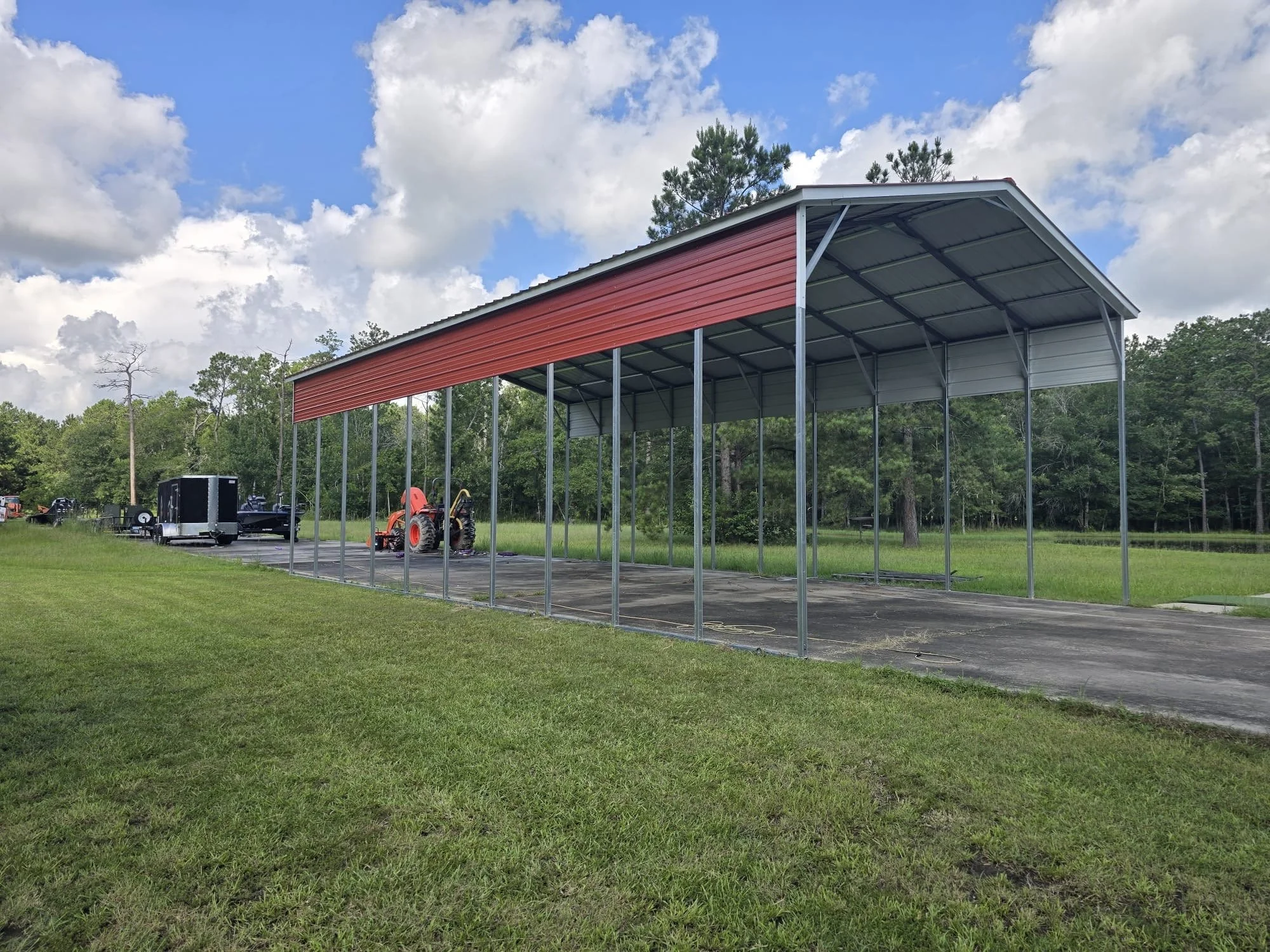 A large metal carport structure with a red roof, situated on a paved area, with a tractor and trailer in the background, surrounded by a grassy field and trees under a partly cloudy sky.