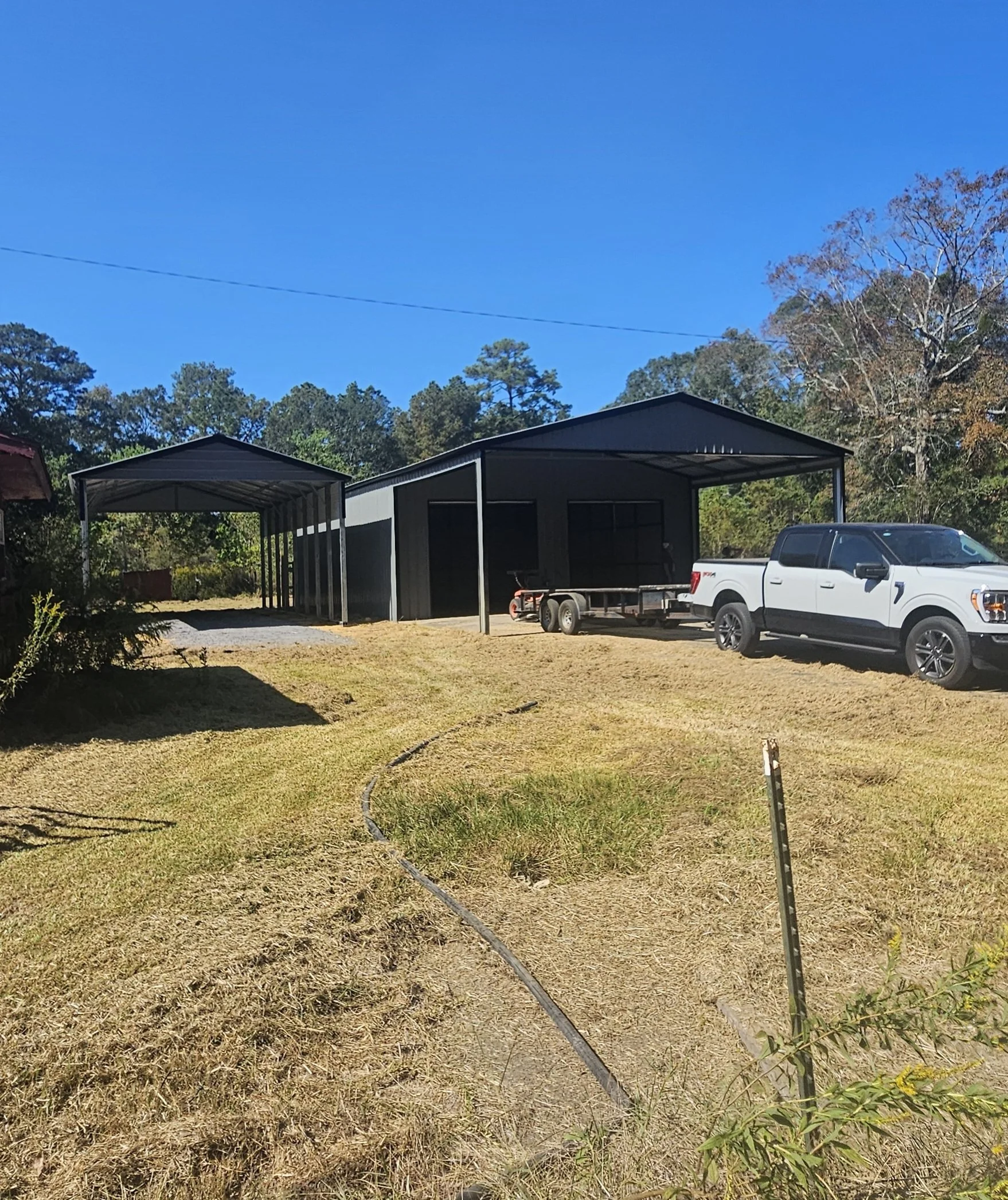 Two metal carports, one with an open front, beside a white pickup truck with a trailer on a gravel driveway, in a grassy yard with trees in the background under a bright blue sky.