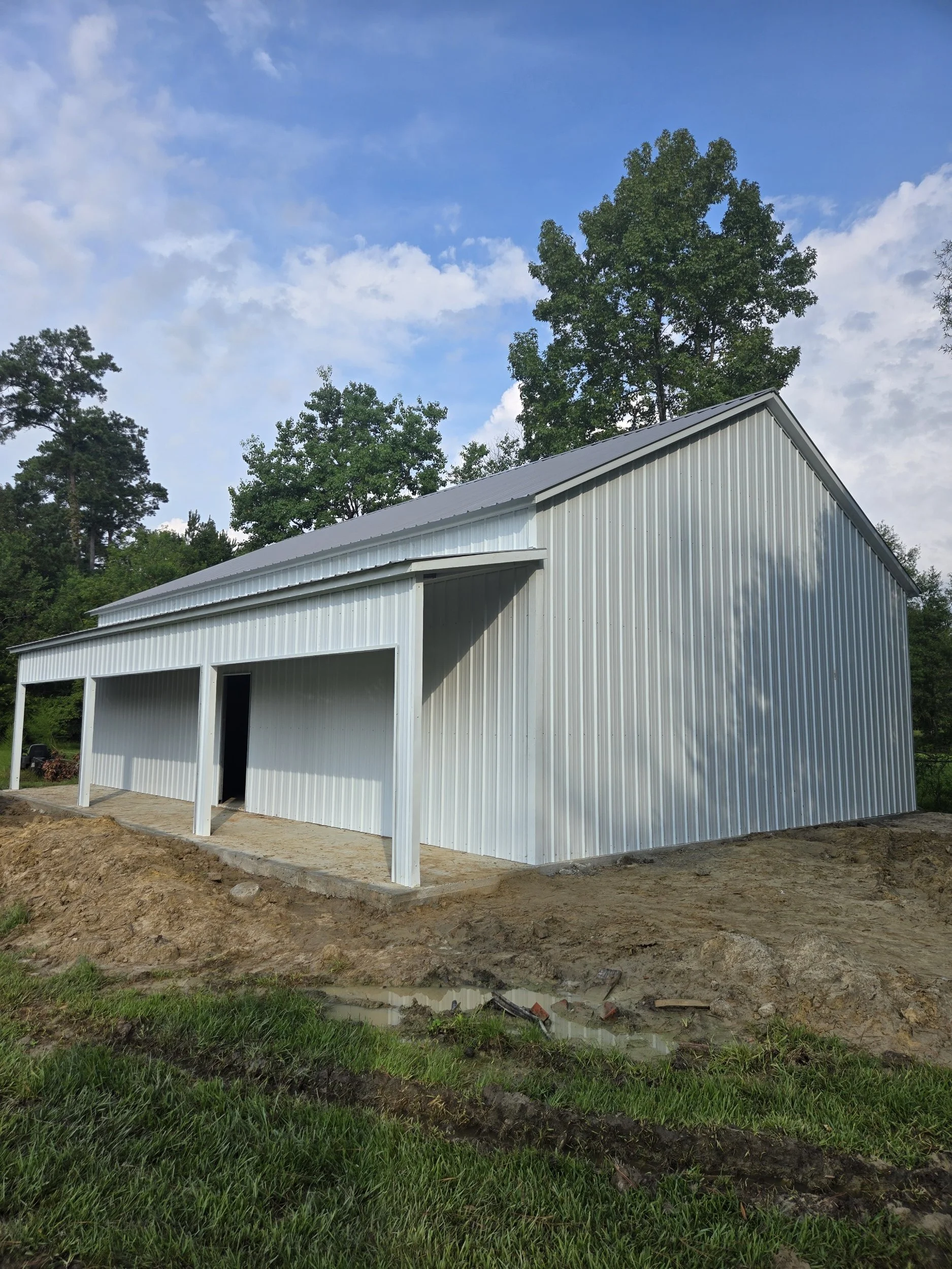 A white metal building, likely a barn or storage shed, with a sloped roof and two open bays, set against a backdrop of tall green trees and a partly cloudy sky.