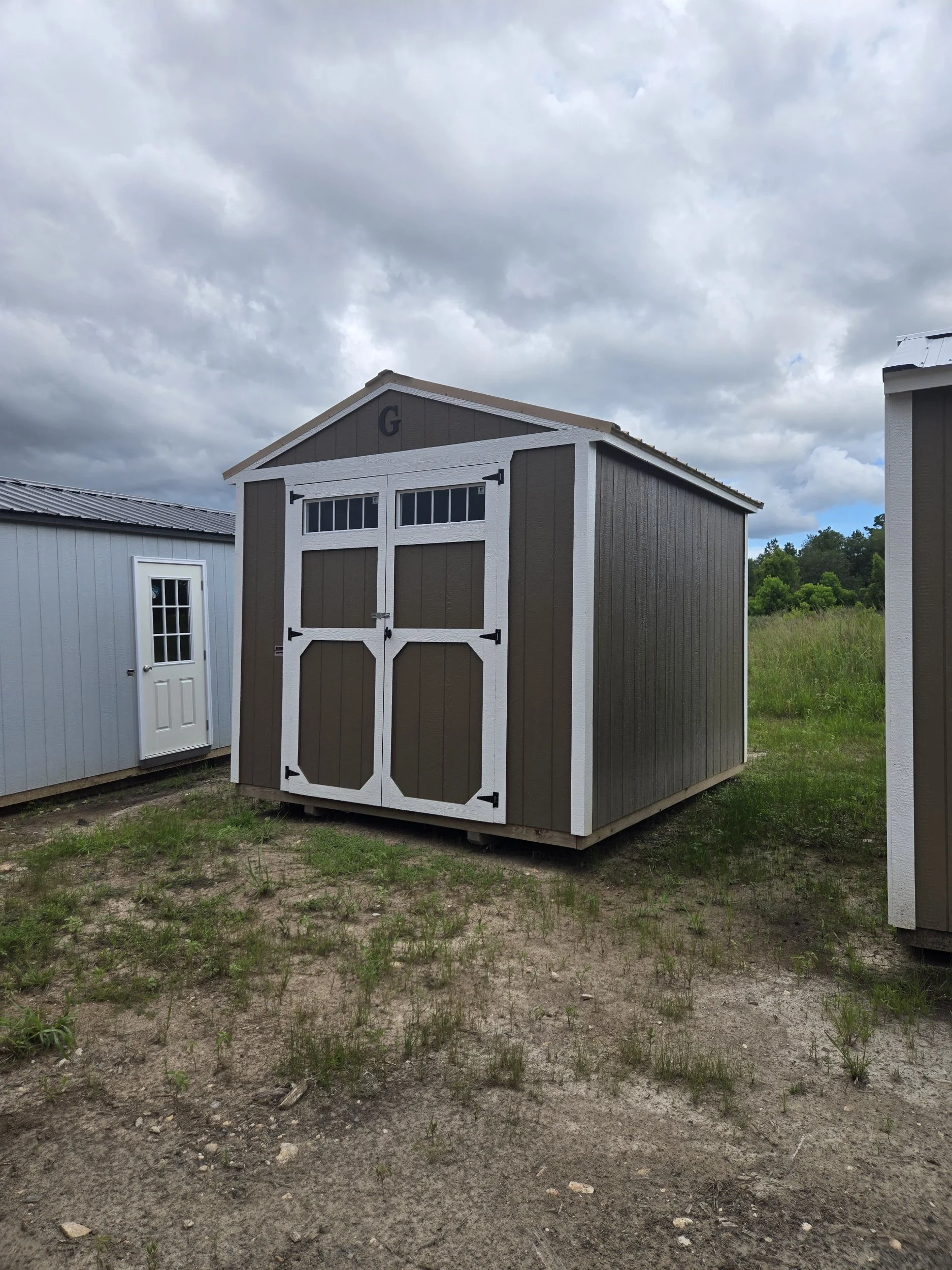 A tan and brown shed with double doors and small windows at the top, situated on a patchy grassy area under a cloudy sky.